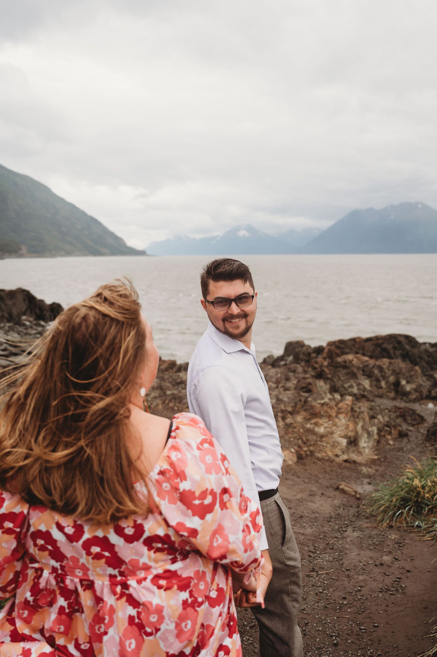 Couple enjoying a scenic moment on rocky shoreline with mountains and water in background on overcast day.