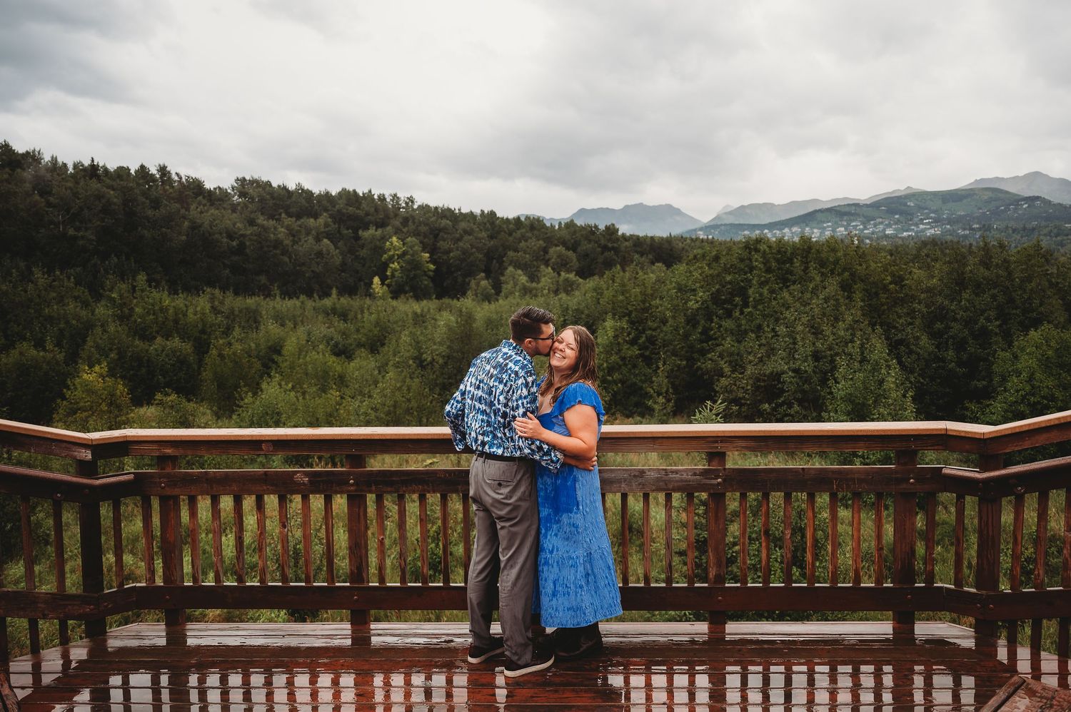 Couple embraces on wooden observation deck overlooking forested mountain landscape under cloudy skies.