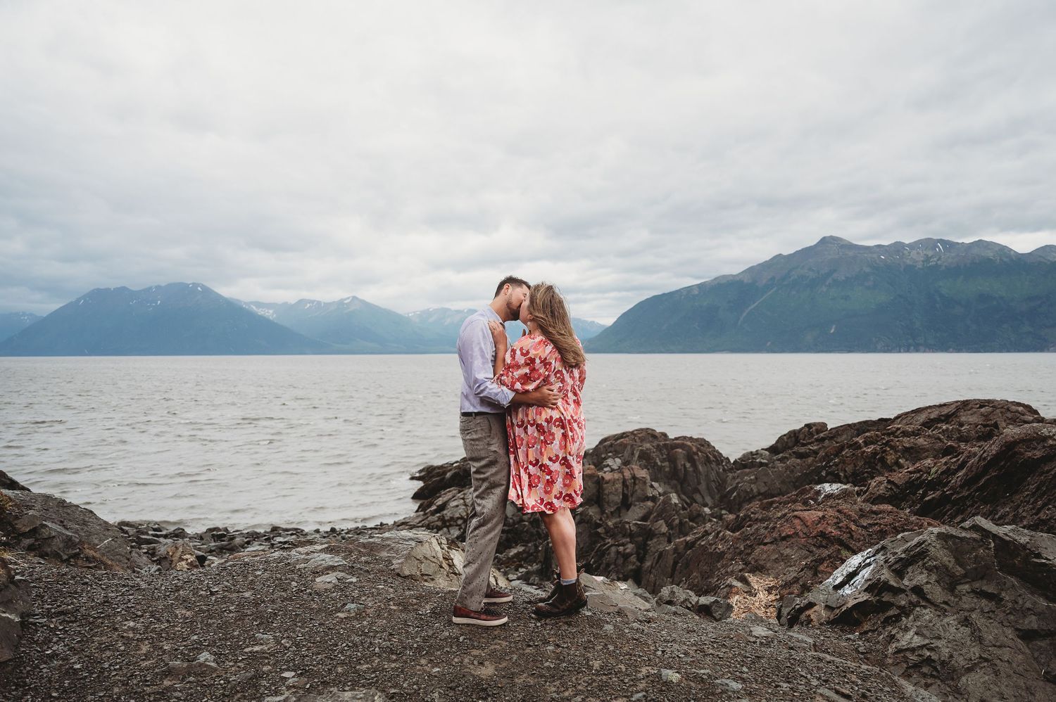 Two people share a romantic moment on a rocky shoreline with mountains and water stretching into the distance.