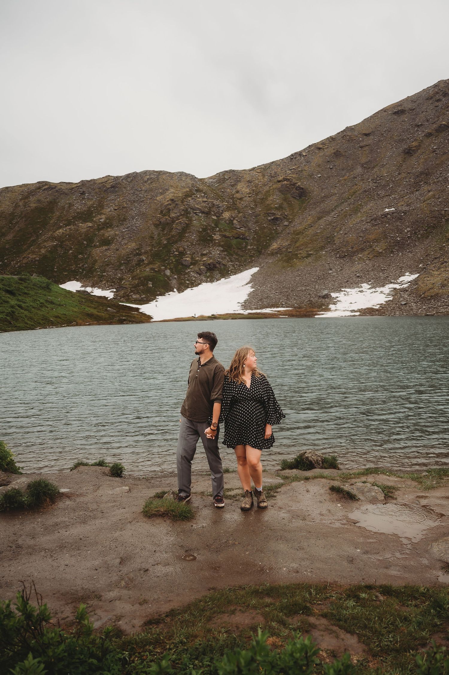 A couple explores a rocky shoreline near an alpine lake surrounded by mountainous terrain.
