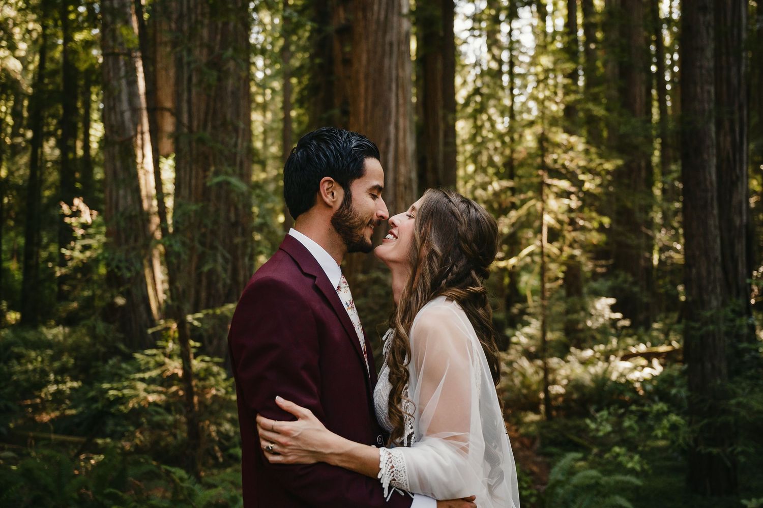Romantic couple in burgundy and white attire share an intimate moment in a sunlit forest setting.