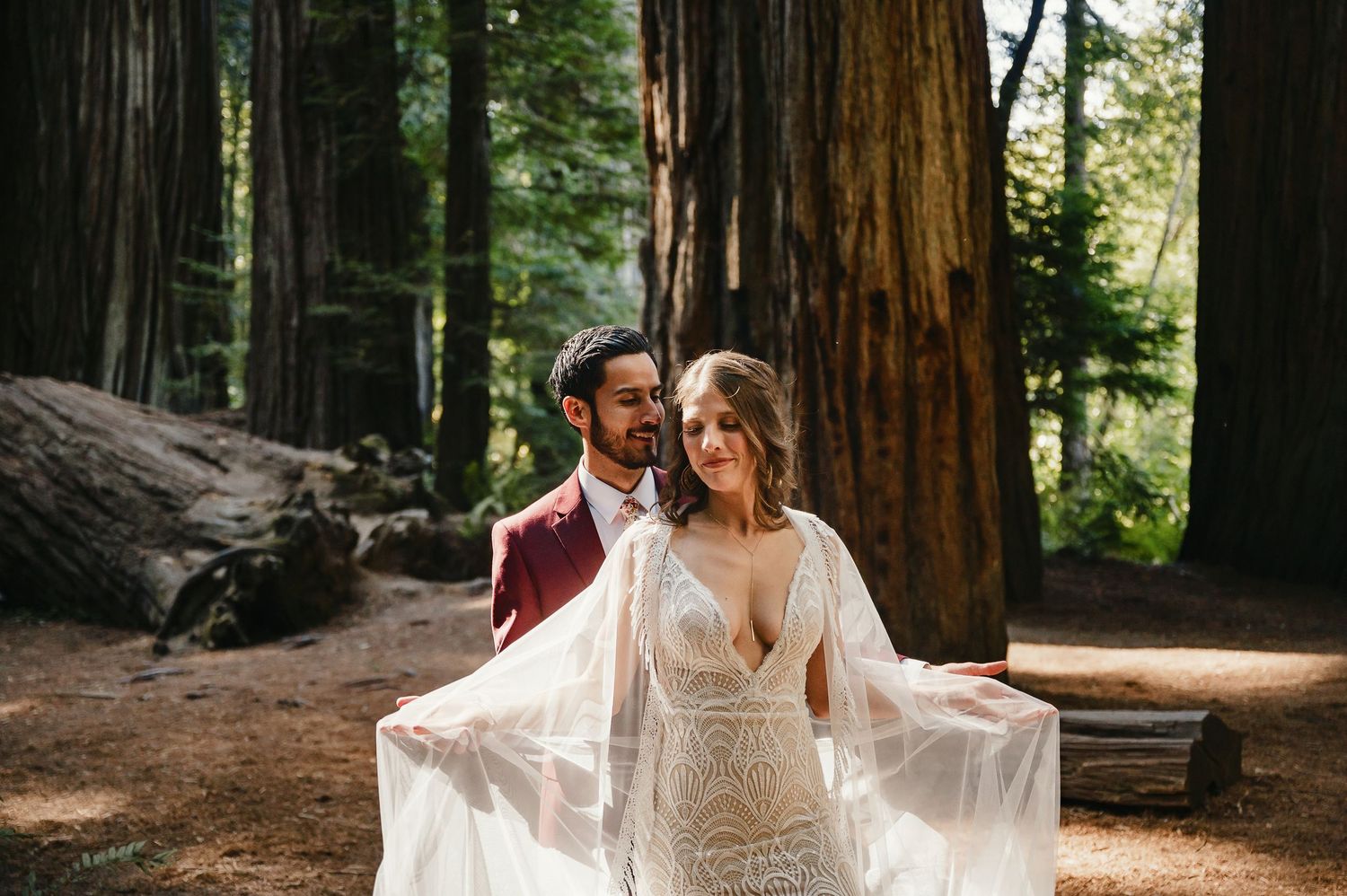 A person in a flowy white wedding dress poses among towering redwood trees in a forest.