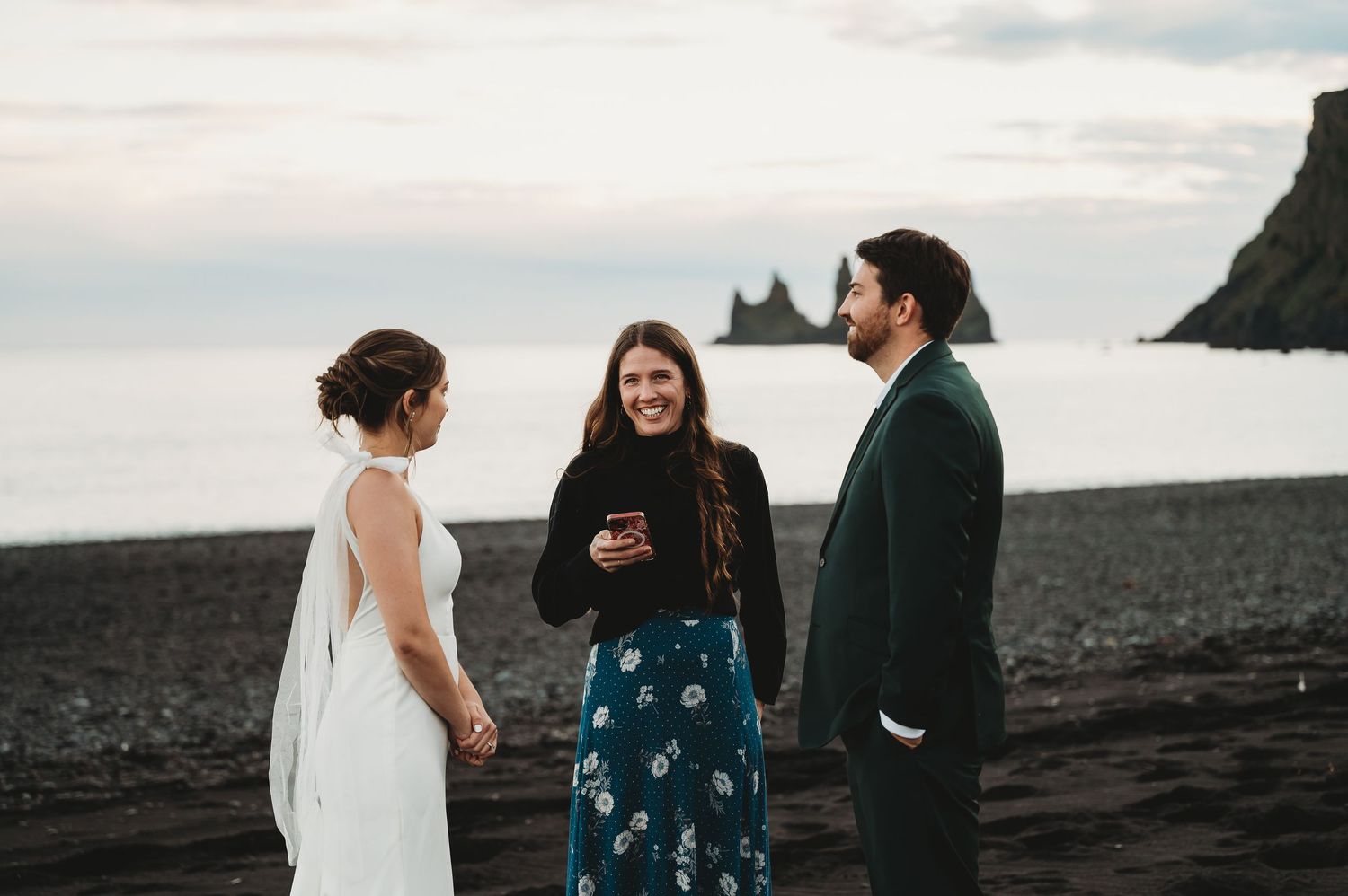 Three people share emotional reactions during a sunset proposal moment on a black sand beach.