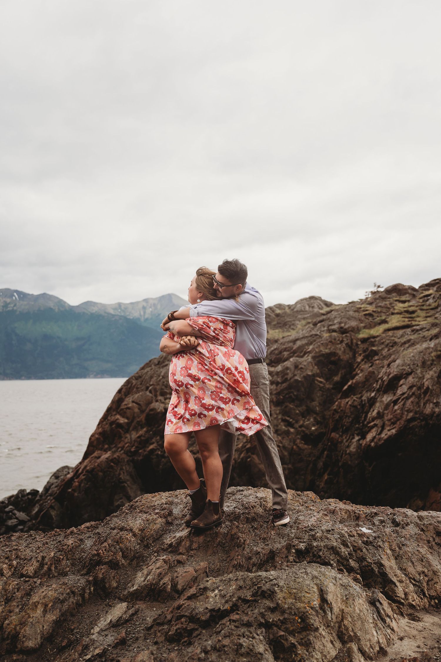Couple sharing intimate moment on rocky coastal cliff overlooking scenic mountain lake vista on overcast day.