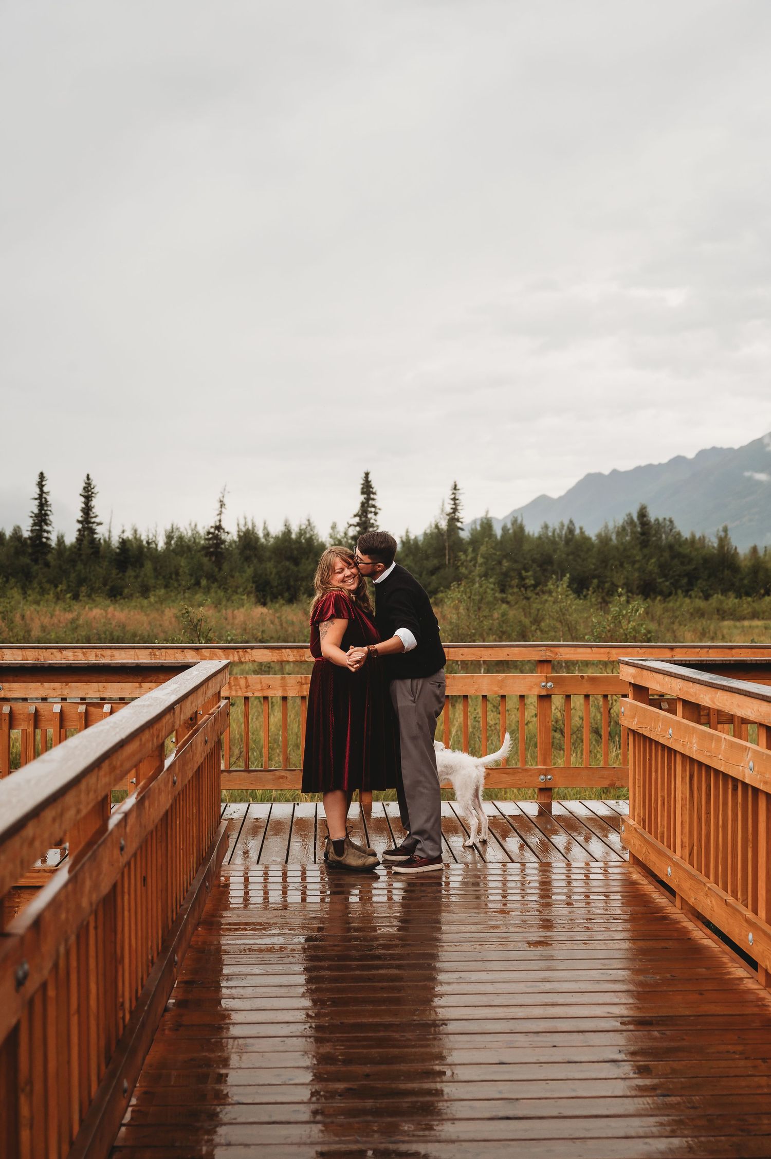 A couple shares a romantic moment on a wet wooden boardwalk with misty mountains in the distance.