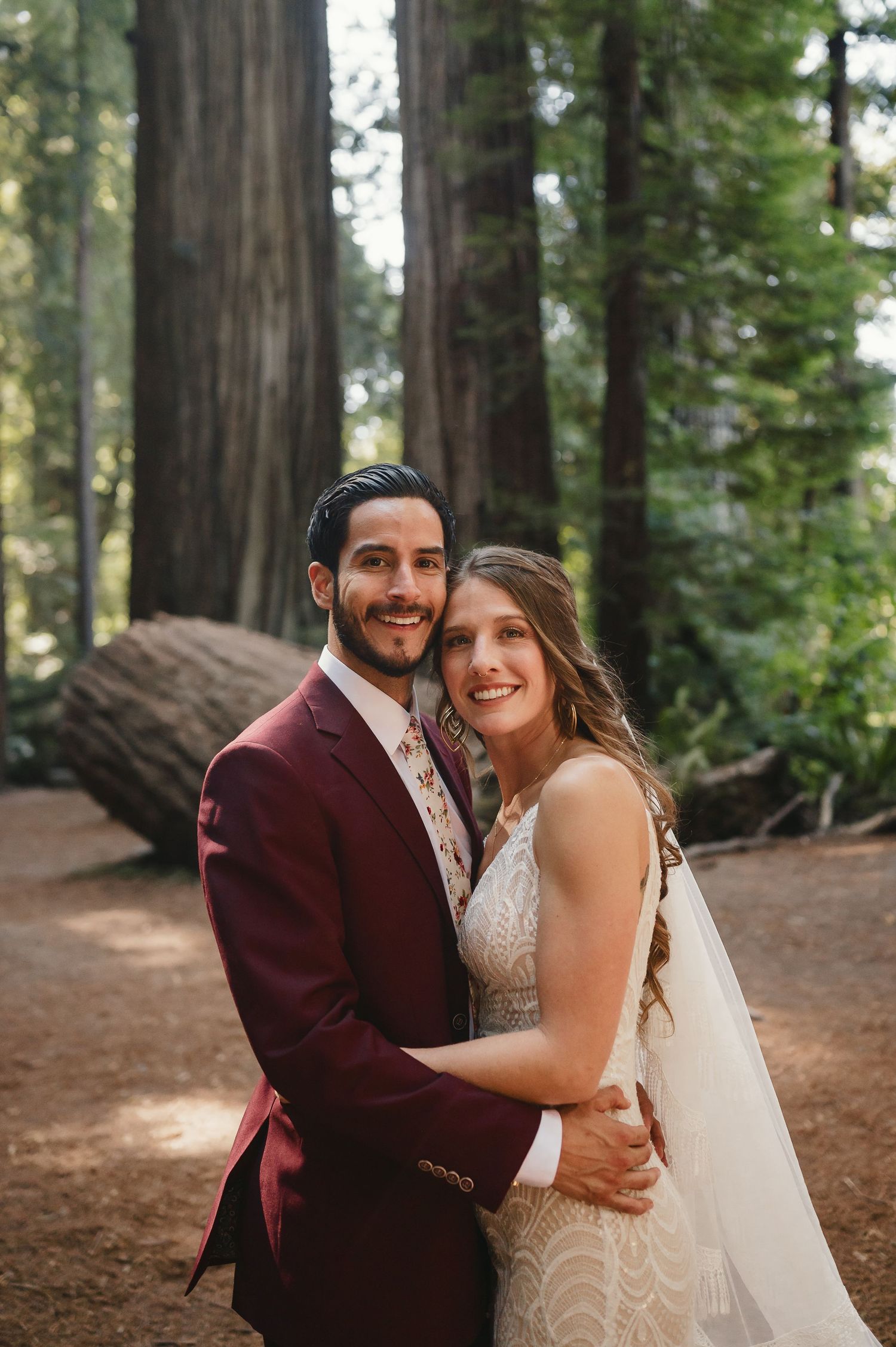 A joyful wedding couple embraces and laughs together in a serene redwood forest setting.