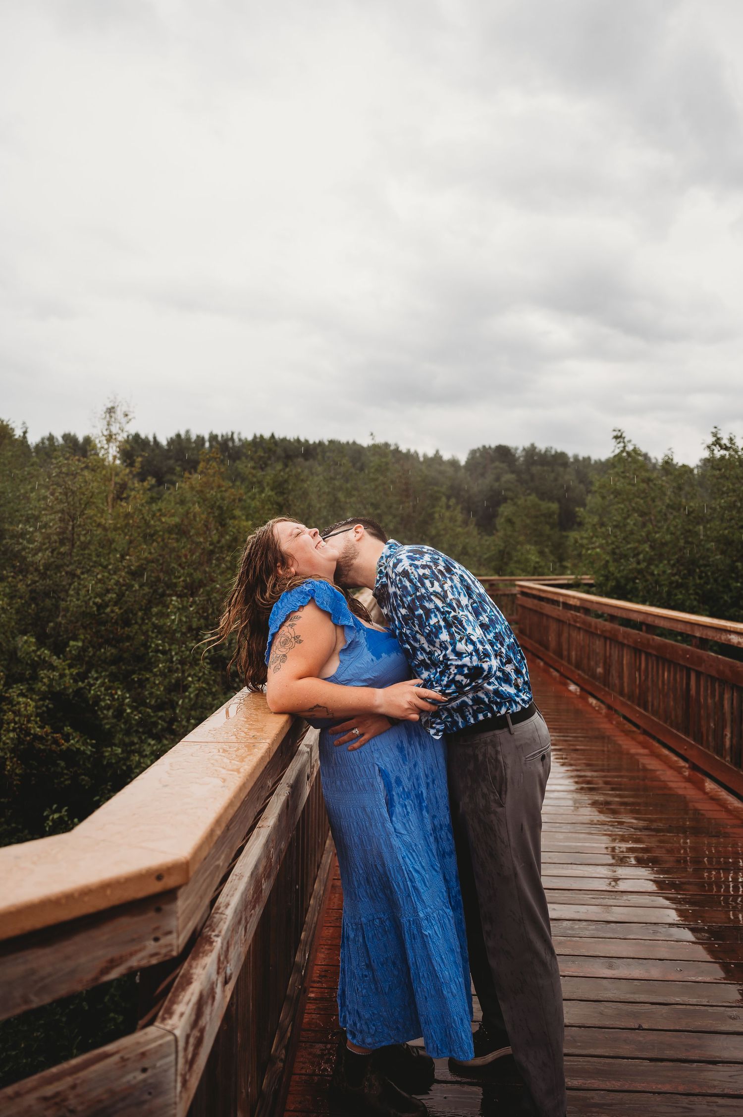 A couple embracing on a wooden bridge walkway surrounded by evergreen forest on a cloudy day.