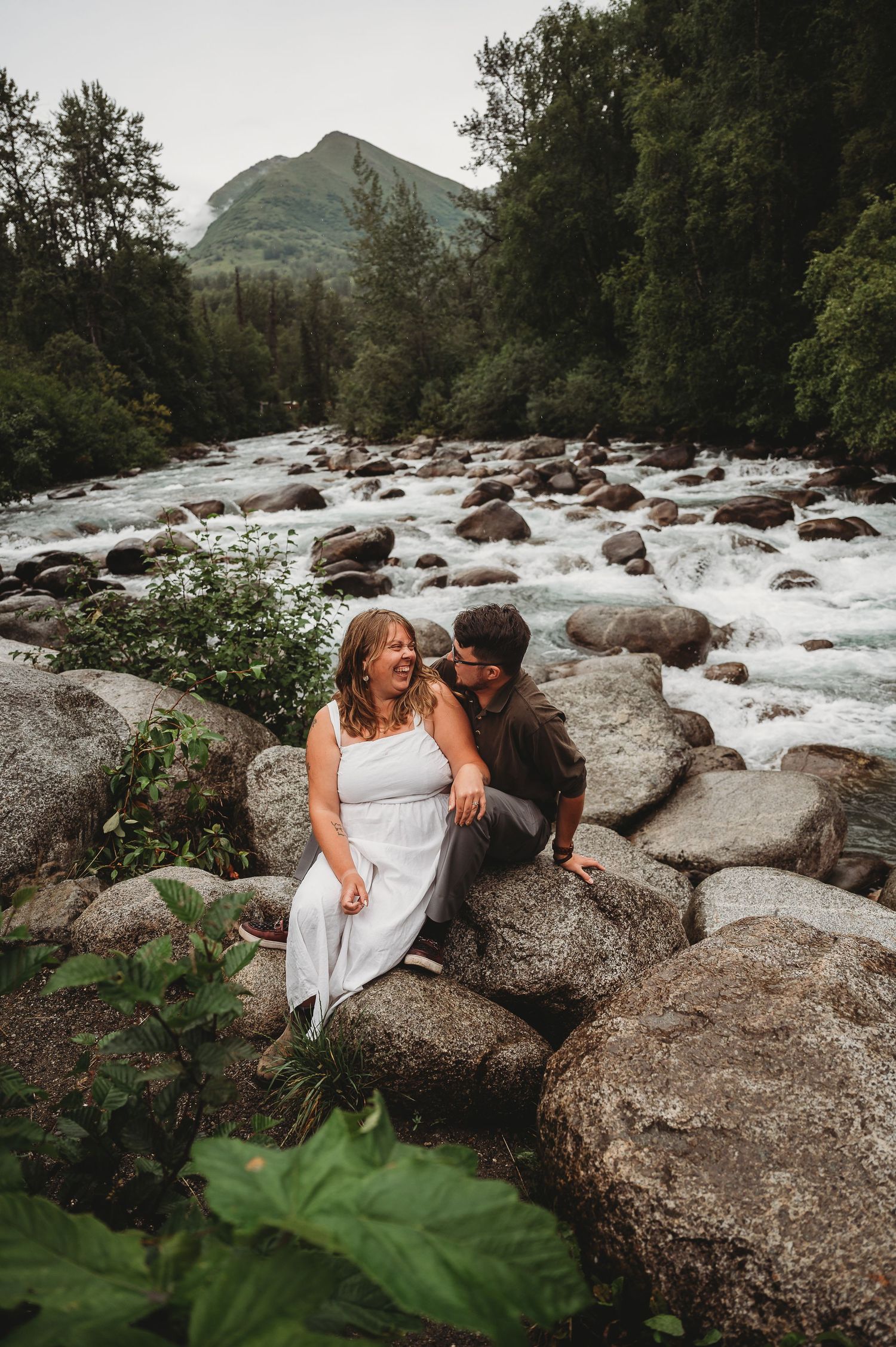 A couple sits together on large boulders next to a rushing mountain stream surrounded by lush greenery.