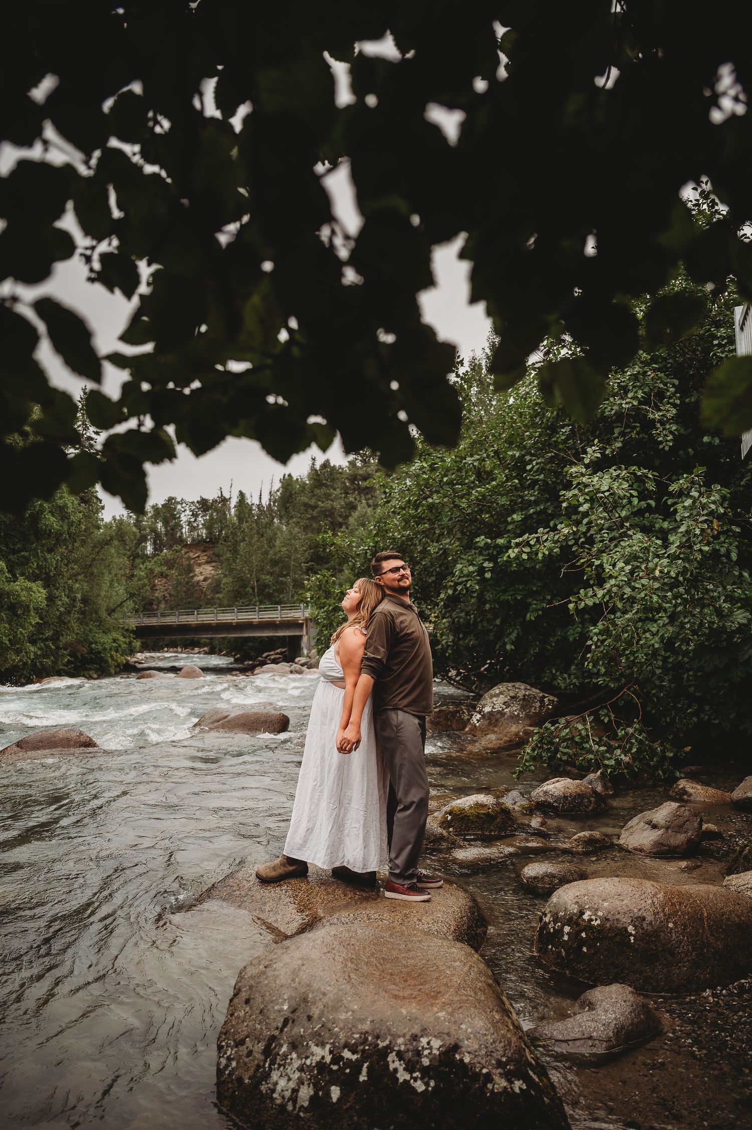 Two figures embrace while standing on large rocks near rushing water in a wooded outdoor setting.