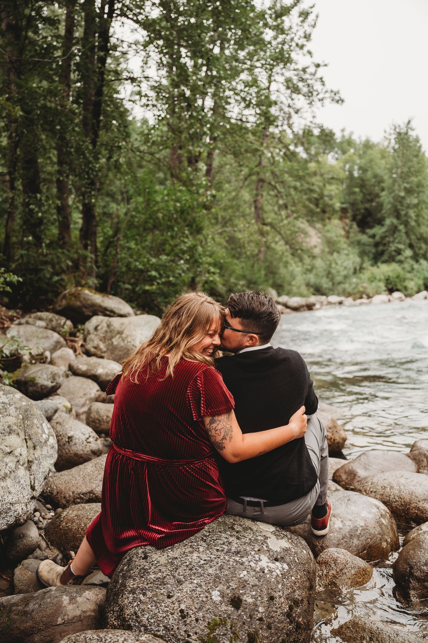 A couple sits together on rocky riverbank surrounded by forest.