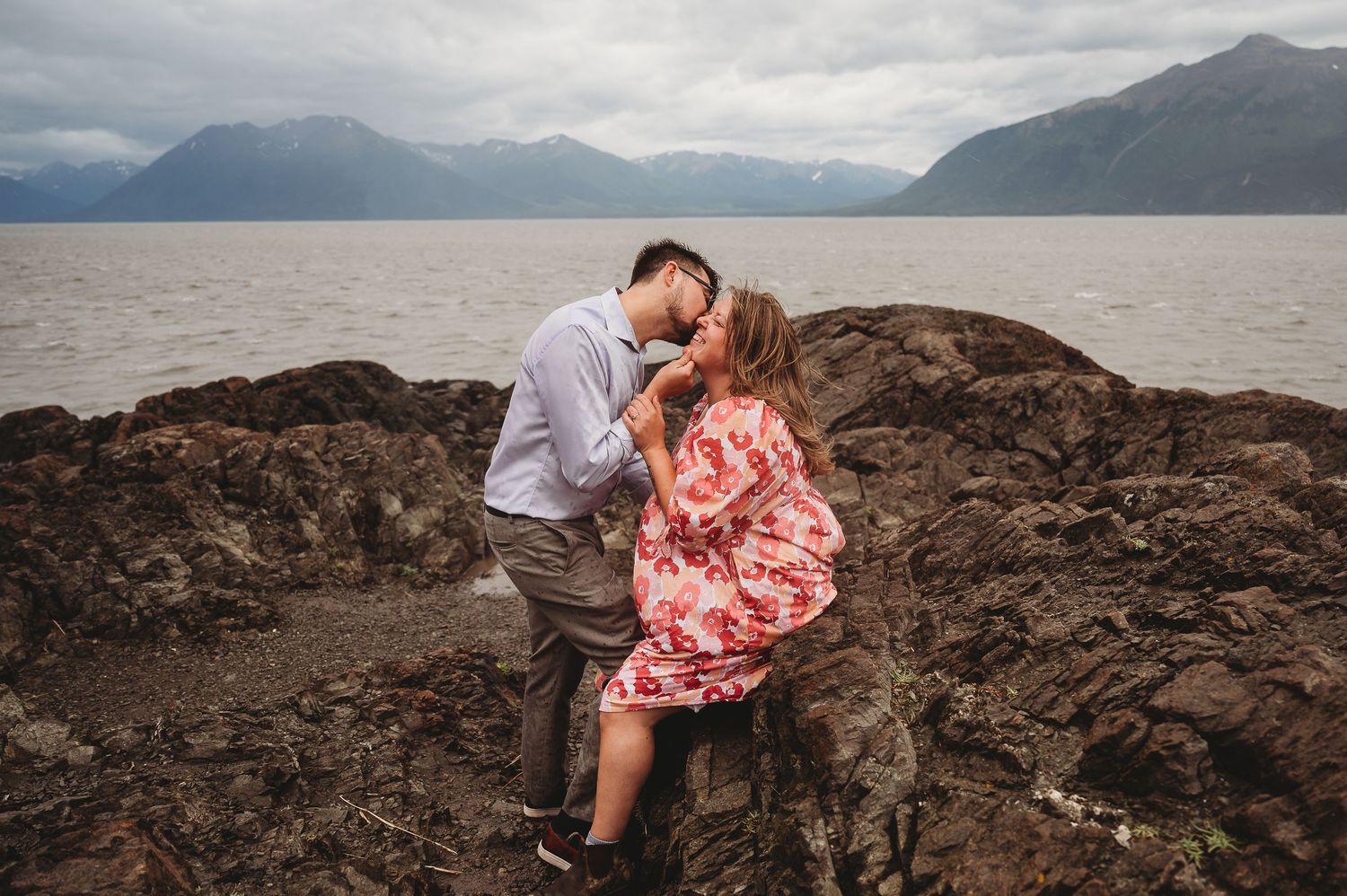 Couple takes romantic engagement photos against dramatic mountain landscape with a cloudy sky and lake backdrop.