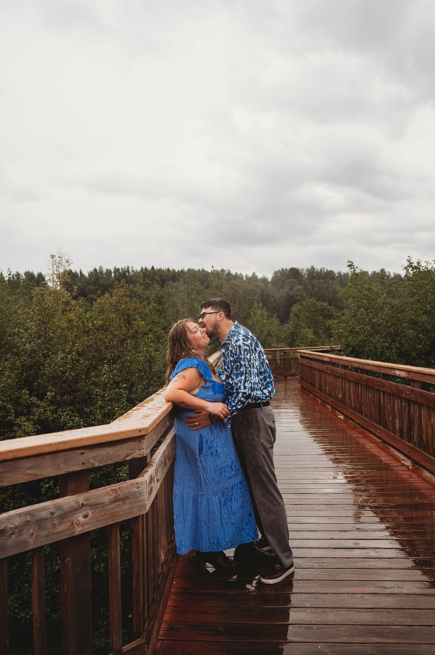 A romantic couple embrace and kiss on a wooden boardwalk surrounded by lush forest on a cloudy day.