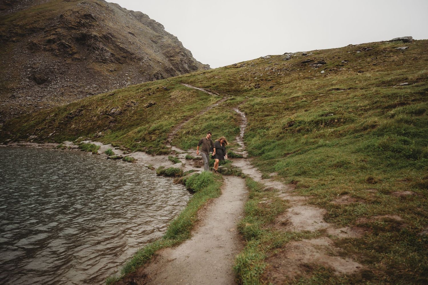 A winding dirt hiking trail leads through rugged mountain terrain under an overcast sky.