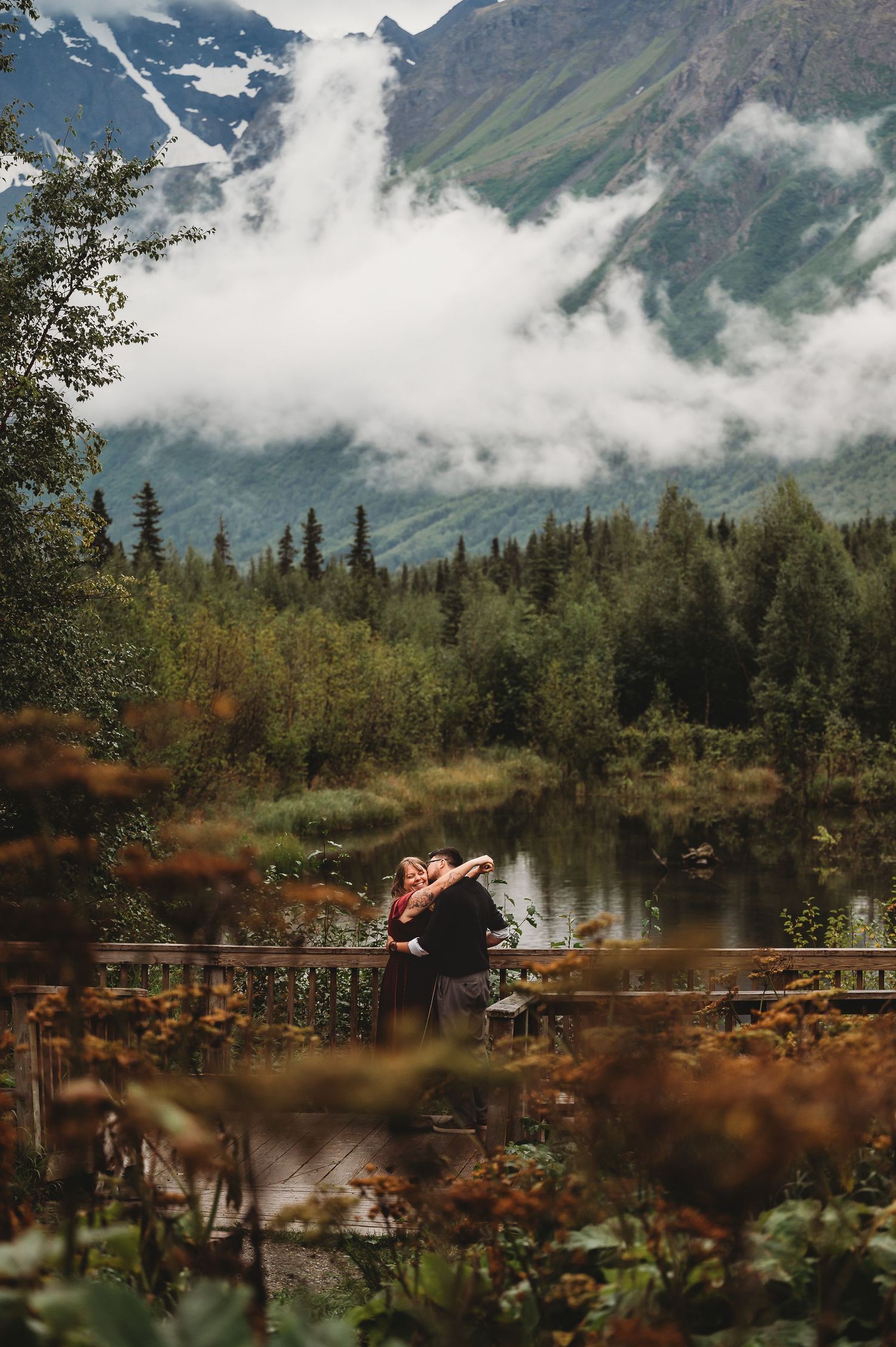 A person sits on a wooden bridge overlooking a mountain lake surrounded by trees and misty peaks.