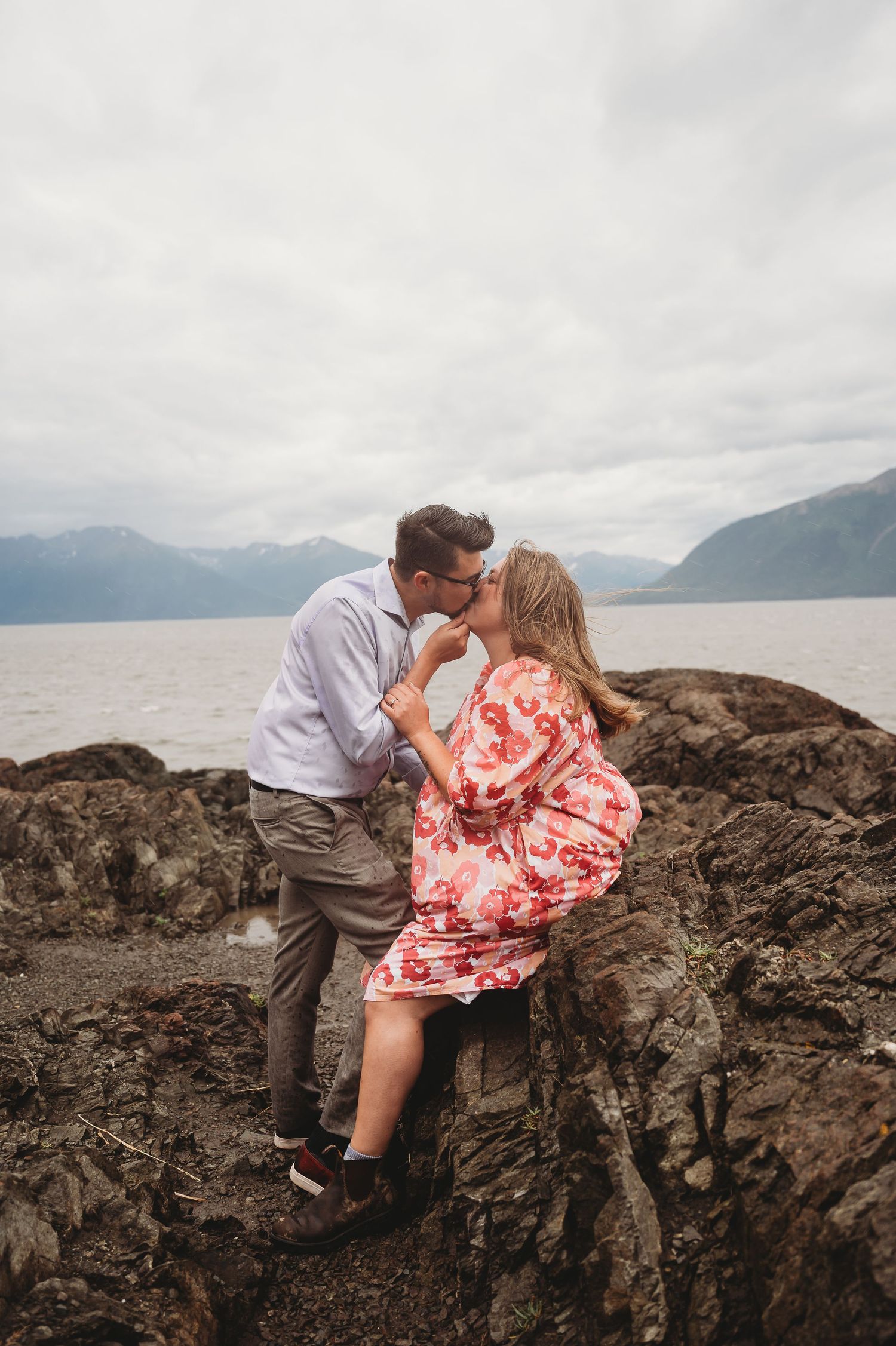 A couple shares a romantic moment on rocky cliffs overlooking a misty mountain lake on an overcast day.