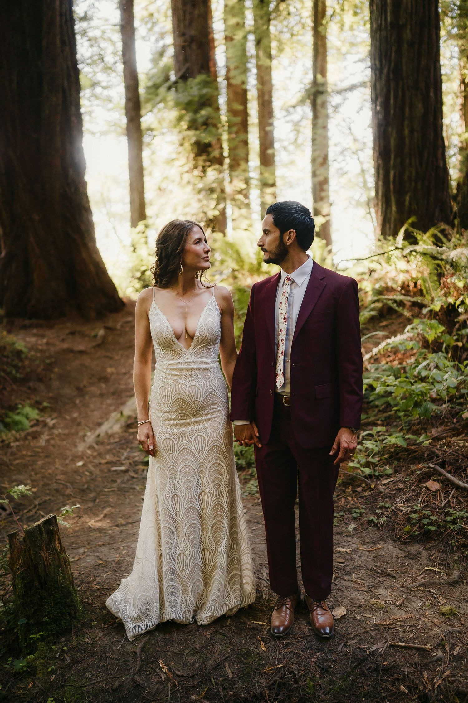 Couple in wedding attire standing together on a forest path surrounded by majestic redwood trees.