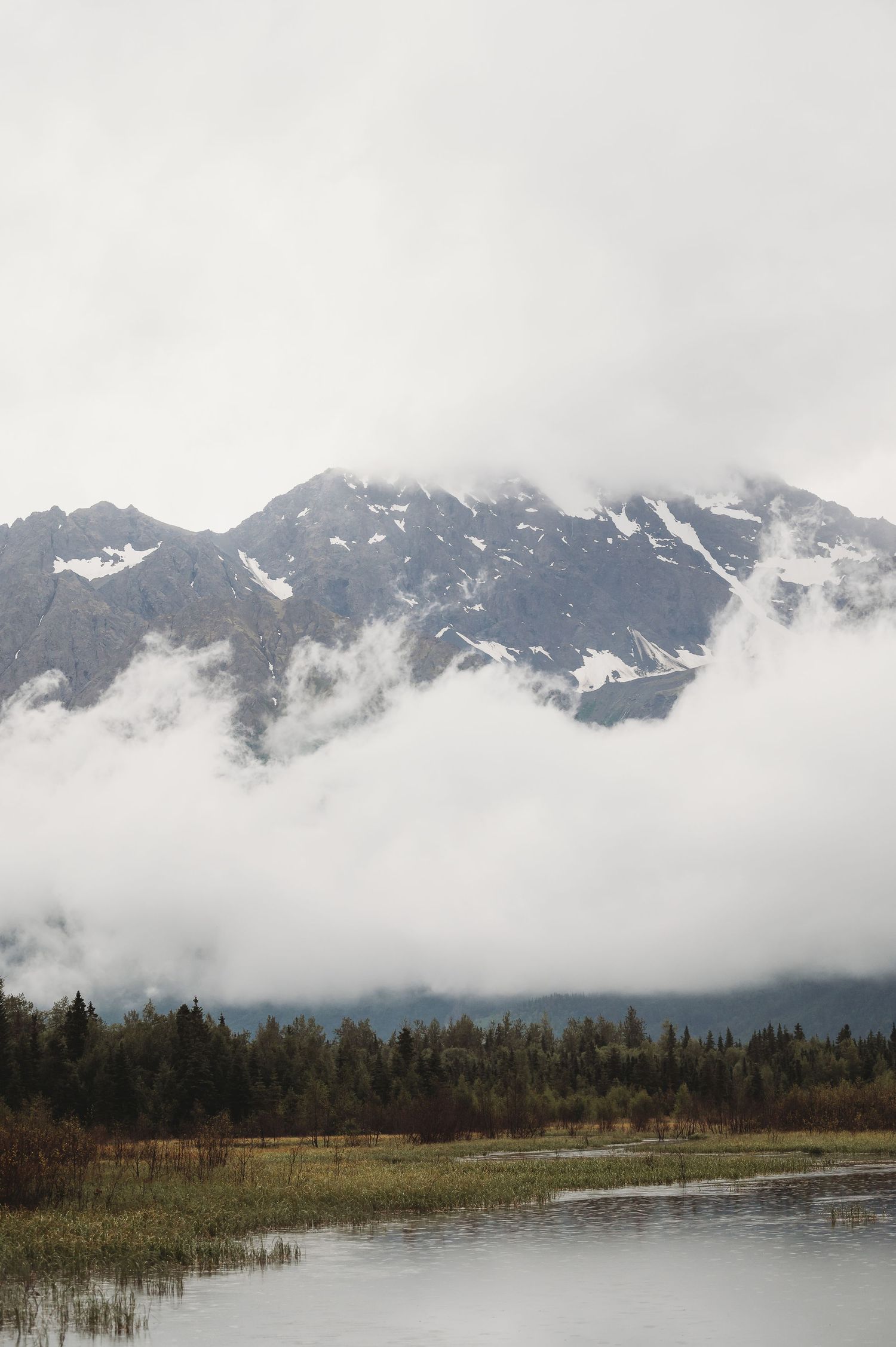 Snow-capped mountains shrouded in fog rise above a marshy lake landscape in Alaska.