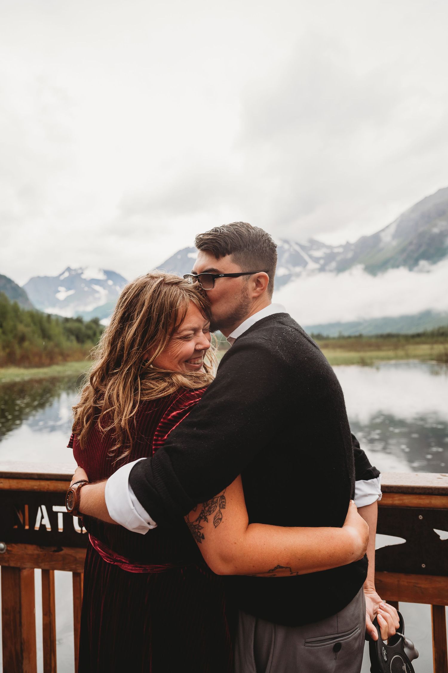 Couple embraces intimately on a wooden deck overlooking misty mountain peaks and a peaceful lake in Alaska.