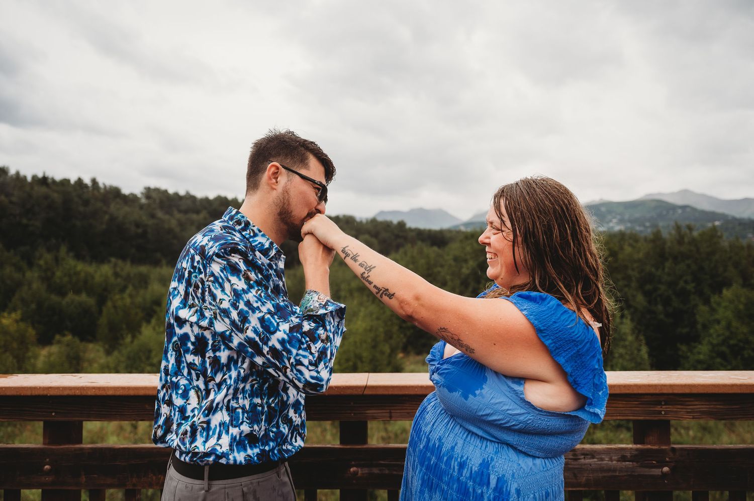 A couple playfully interacts on an outdoor deck overlooking a forest and mountain landscape in the background.