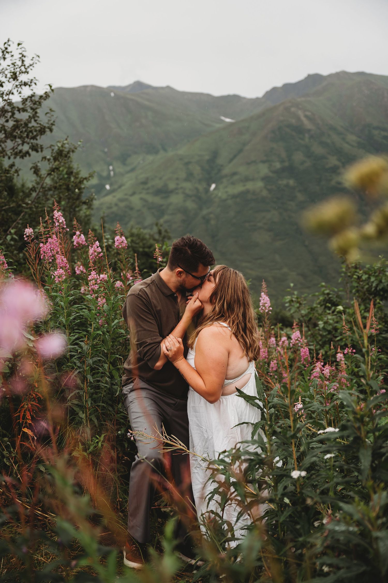 A couple shares a romantic moment among pink wildflowers with mountain peaks in the background.