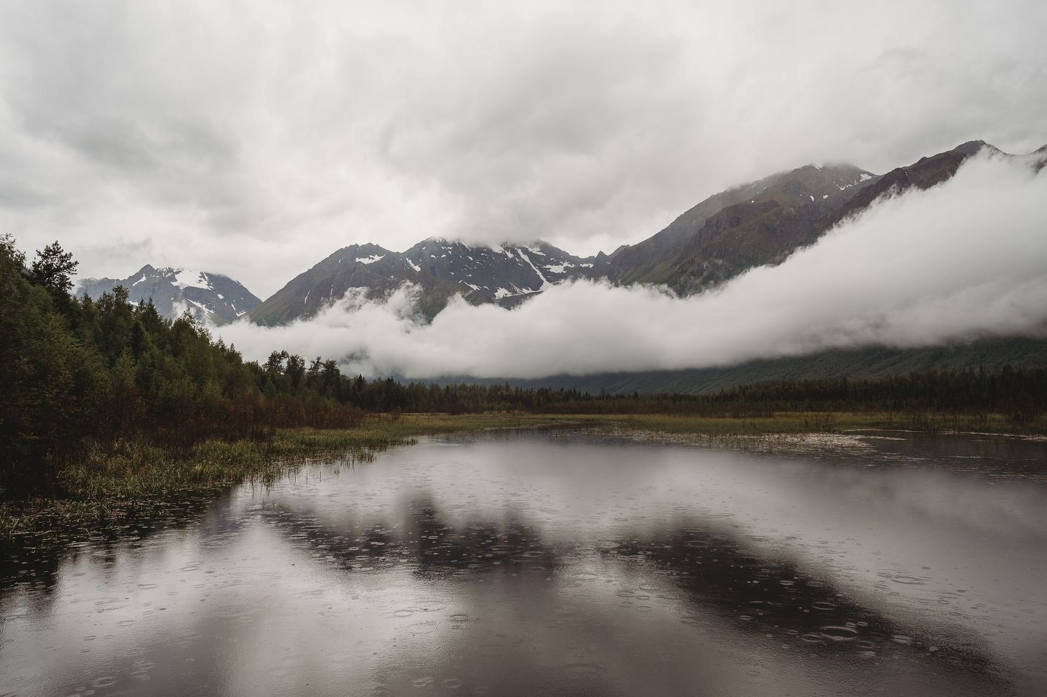 Misty mountains reflect in a still lake surrounded by evergreen forest in a moody landscape.