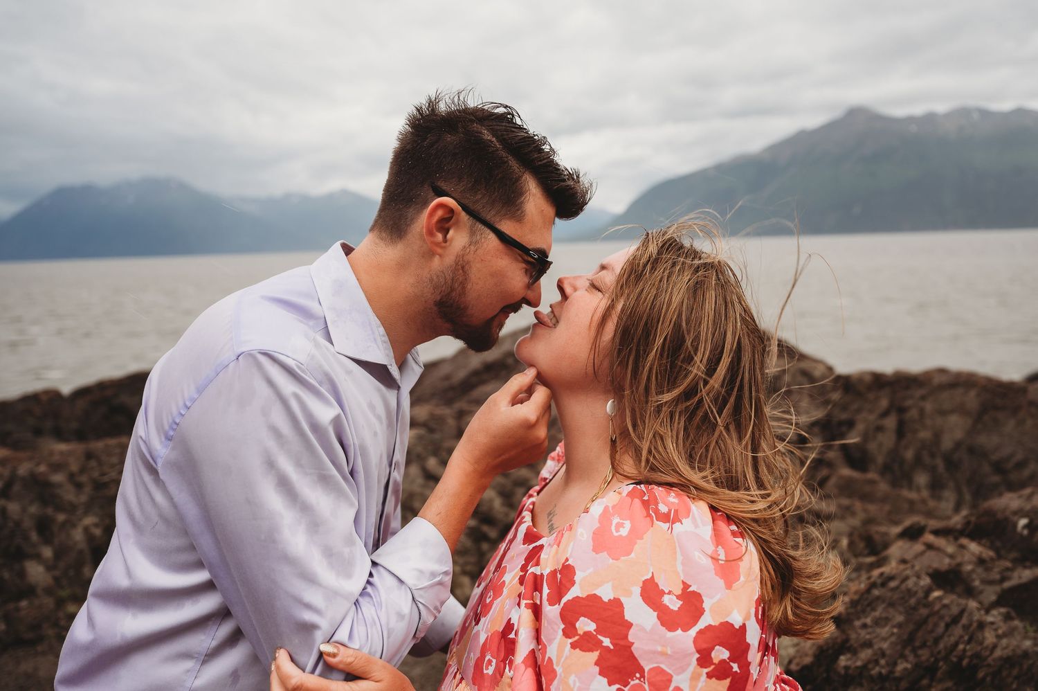 A romantic couple shares an intimate moment near a mountain lake, with the woman wearing a coral patterned dress.