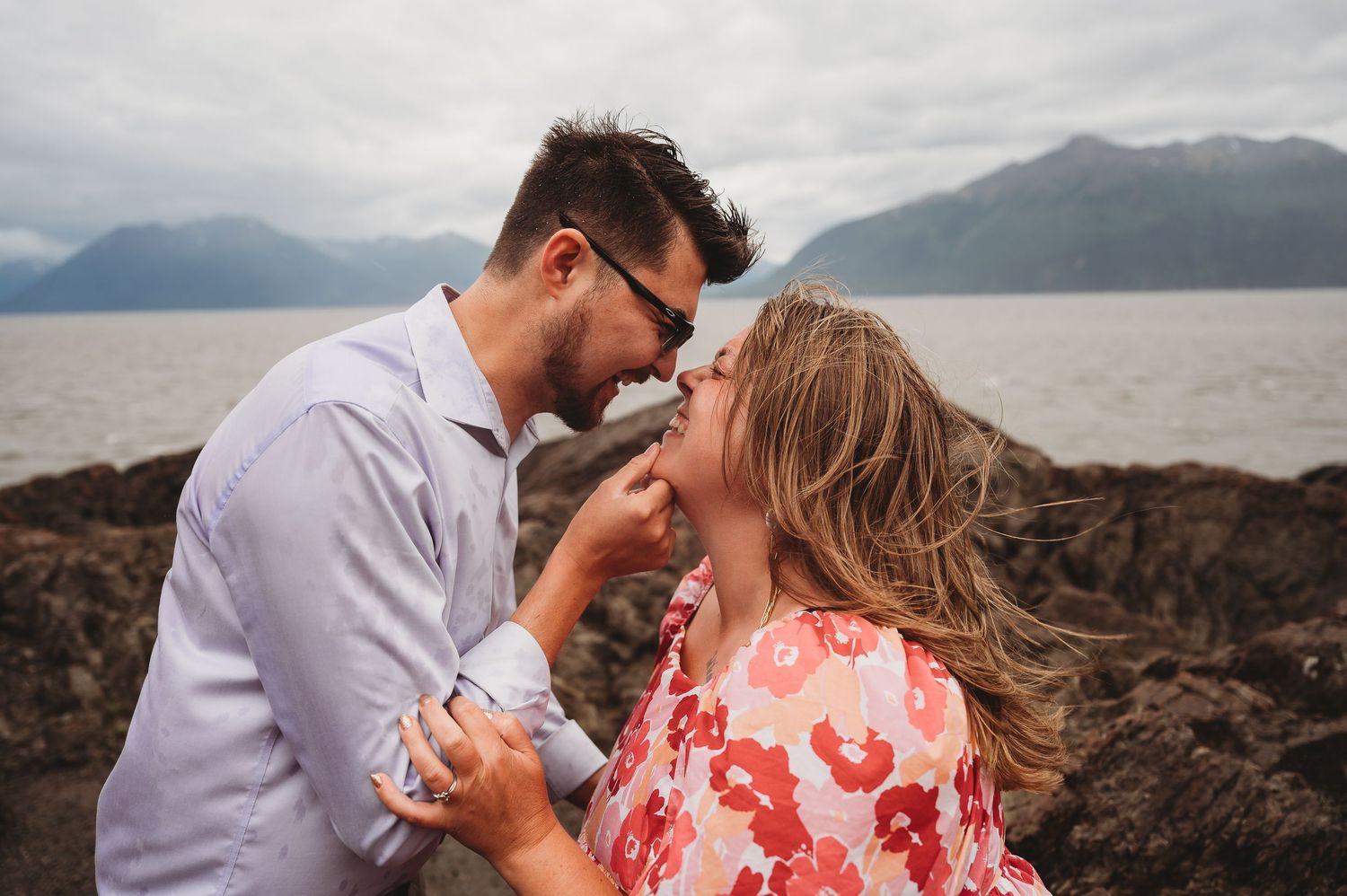 A romantic moment between a couple on rocky coastal cliffs with mountains and water in the background.