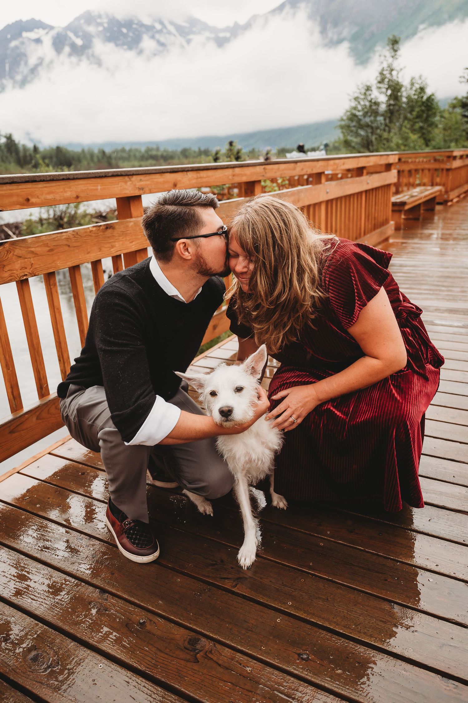 A couple shares a tender moment with their white dog on a wet wooden deck overlooking misty mountain views.