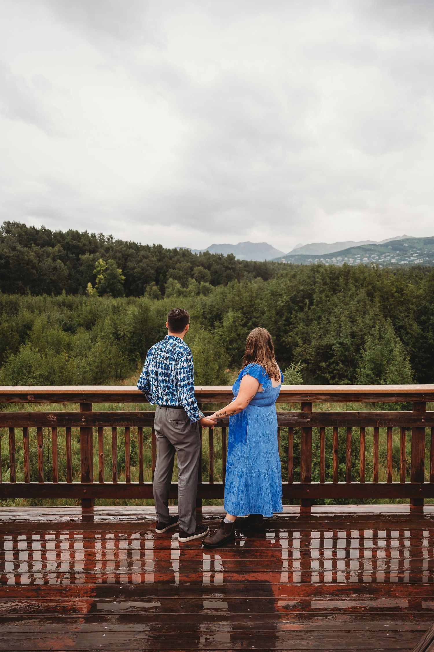 Two people enjoying a scenic view while sitting on a wooden deck railing with forest and mountains in the background.