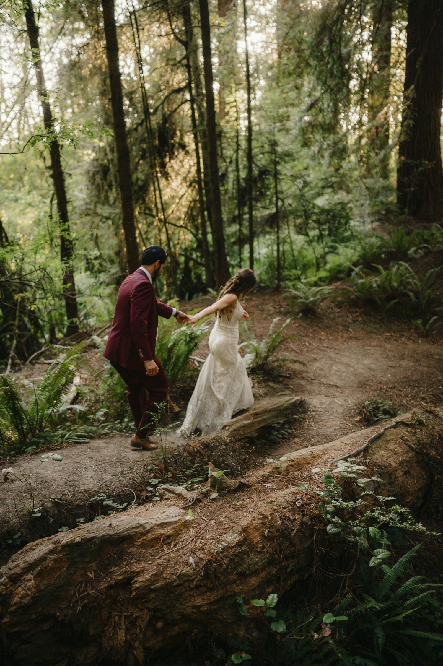 Newlyweds walking hand in hand along a winding forest path surrounded by lush greenery and filtered sunlight.