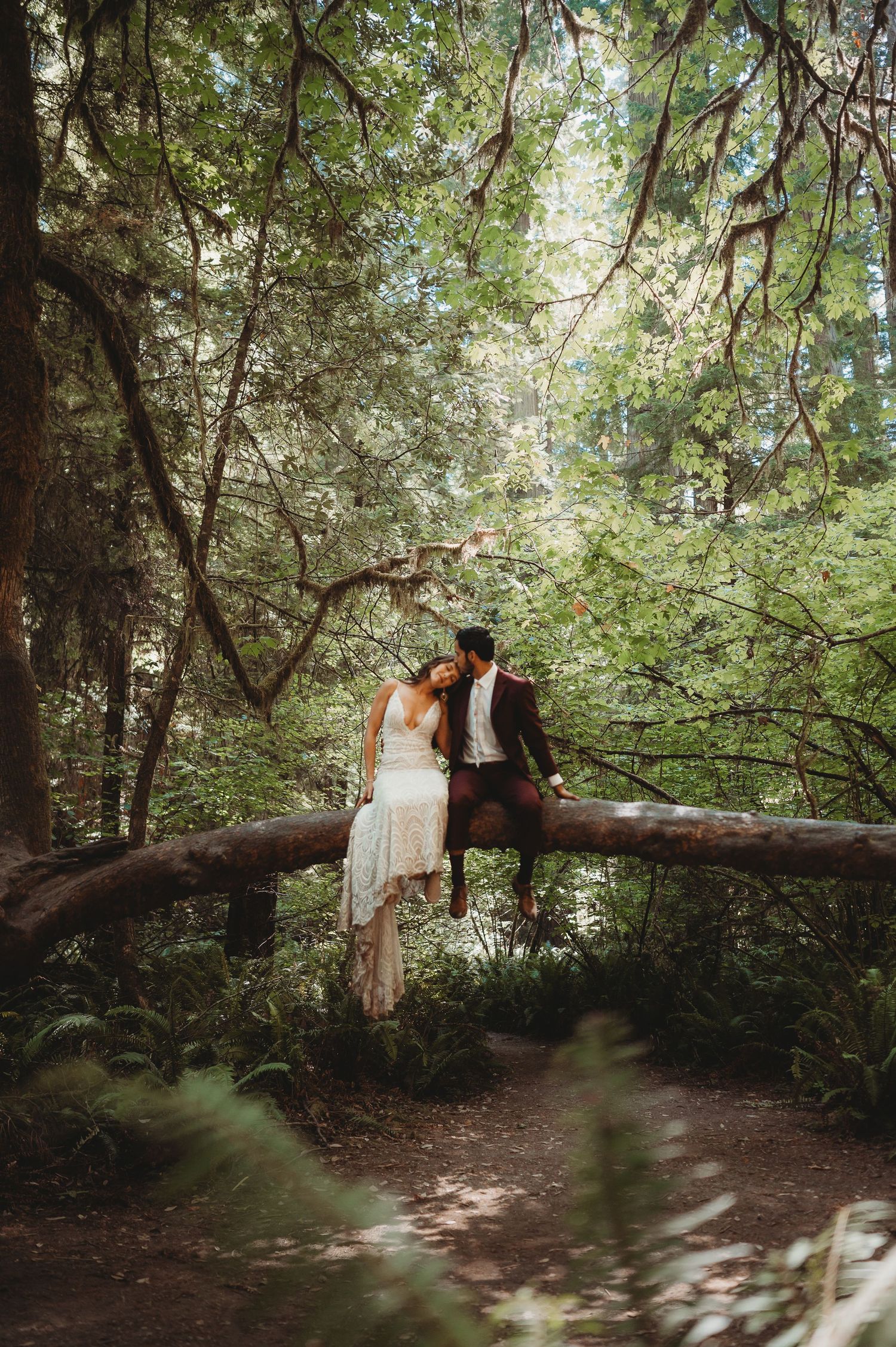 A couple sits together on a fallen log bridge in a misty green forest setting during a romantic photoshoot.