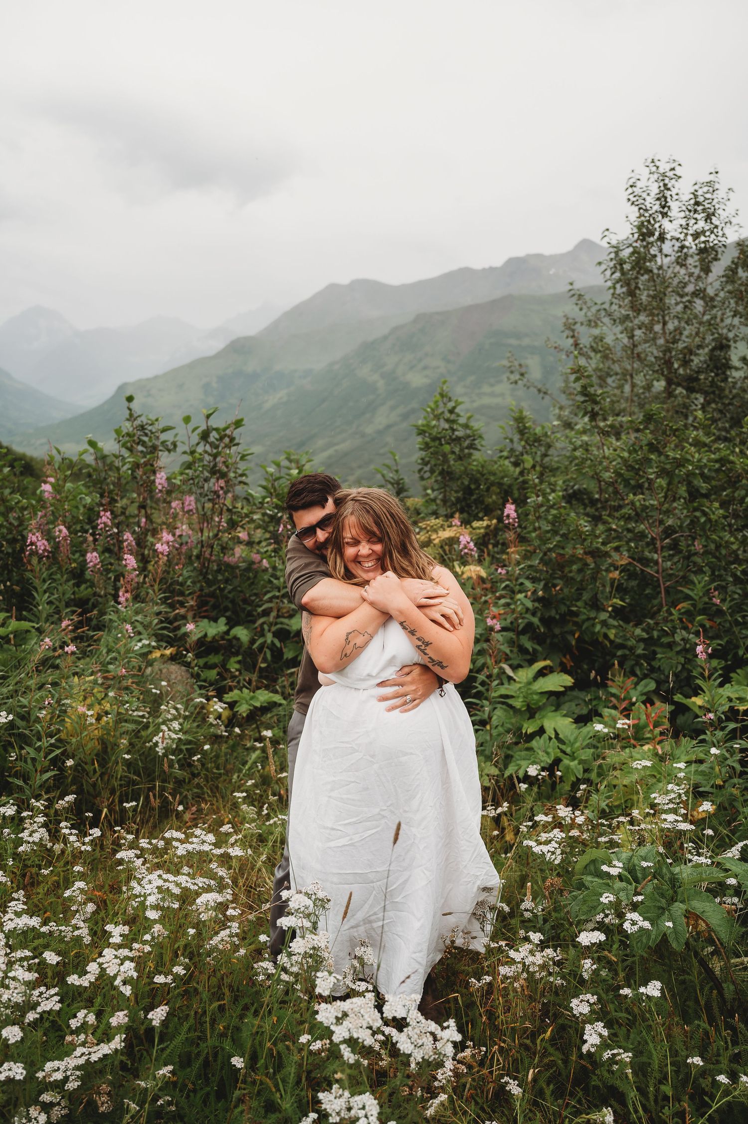 Two figures embrace in a field of wildflowers with misty mountain peaks rising in the background.
