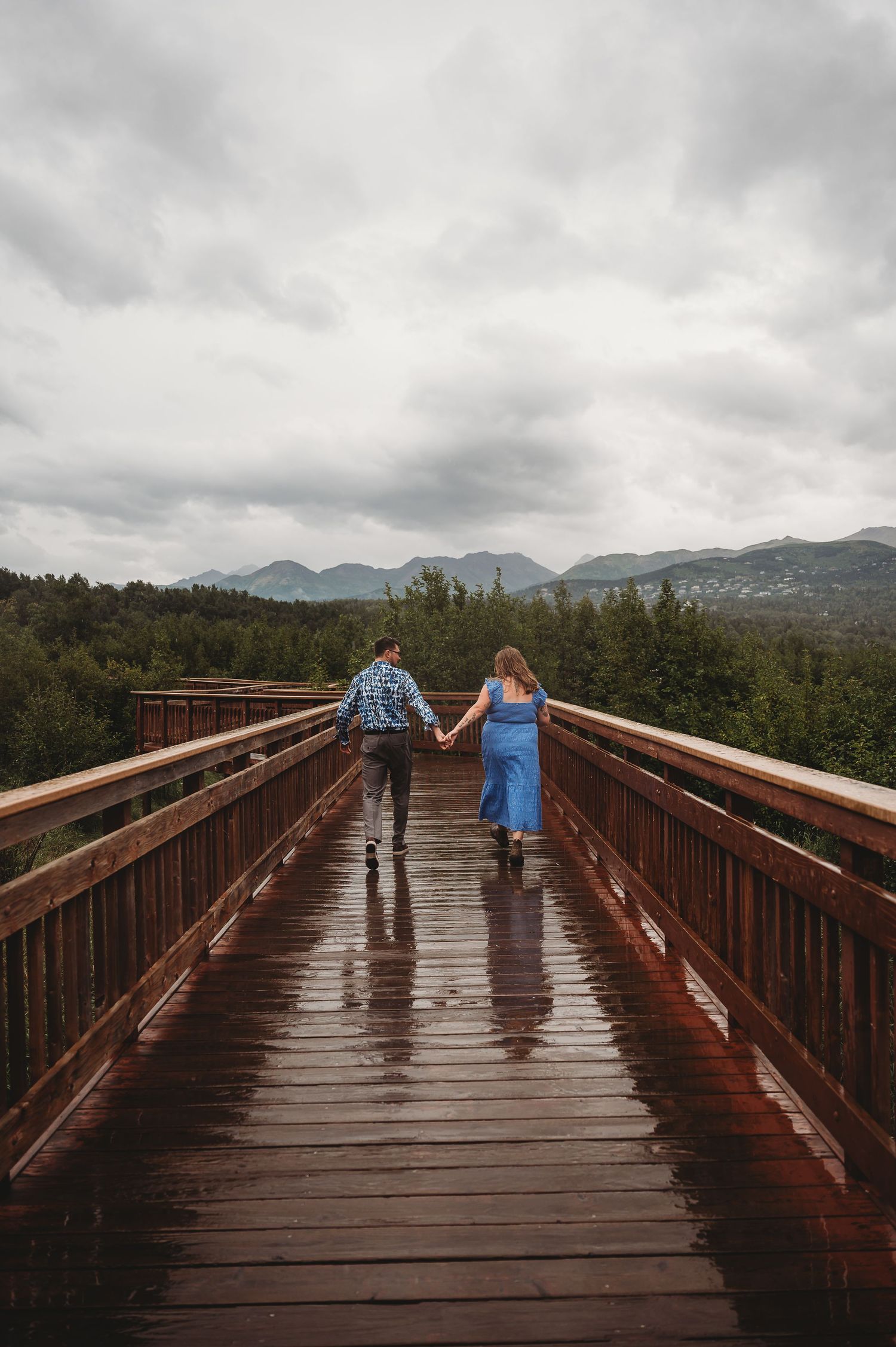 People walking on a wet wooden boardwalk surrounded by mountains and forest on a cloudy rainy day in nature.