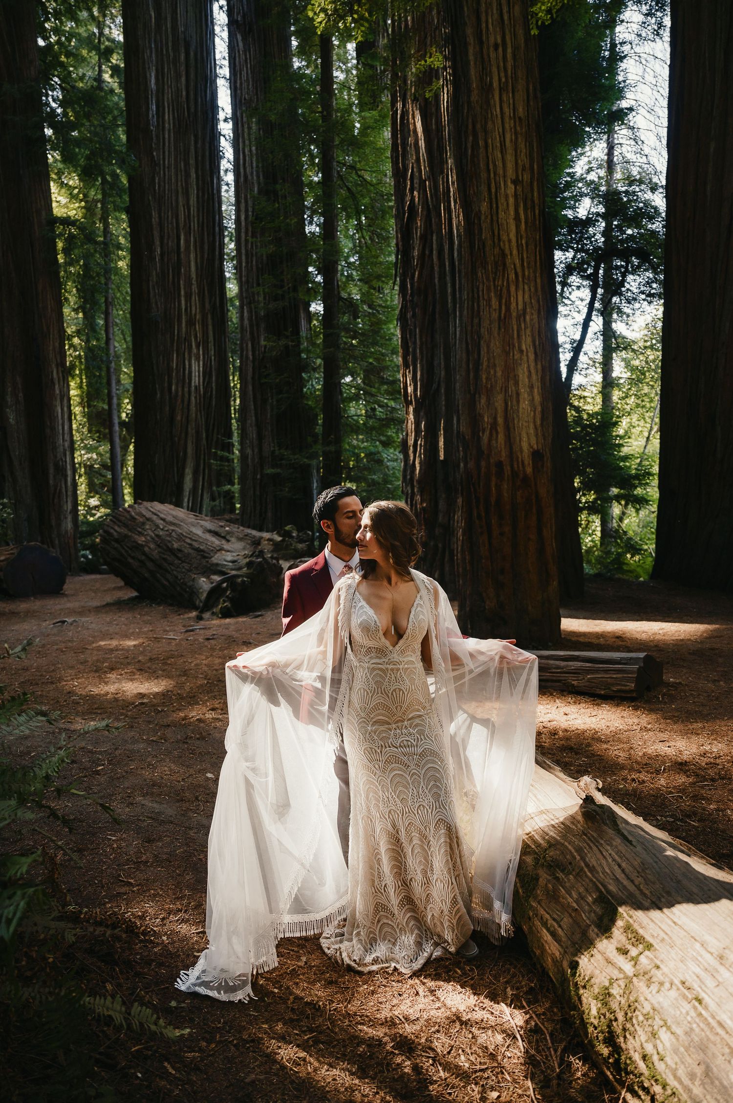 A romantic wedding couple poses in a sunlit redwood forest wearing a flowing lace dress with billowing sleeves.