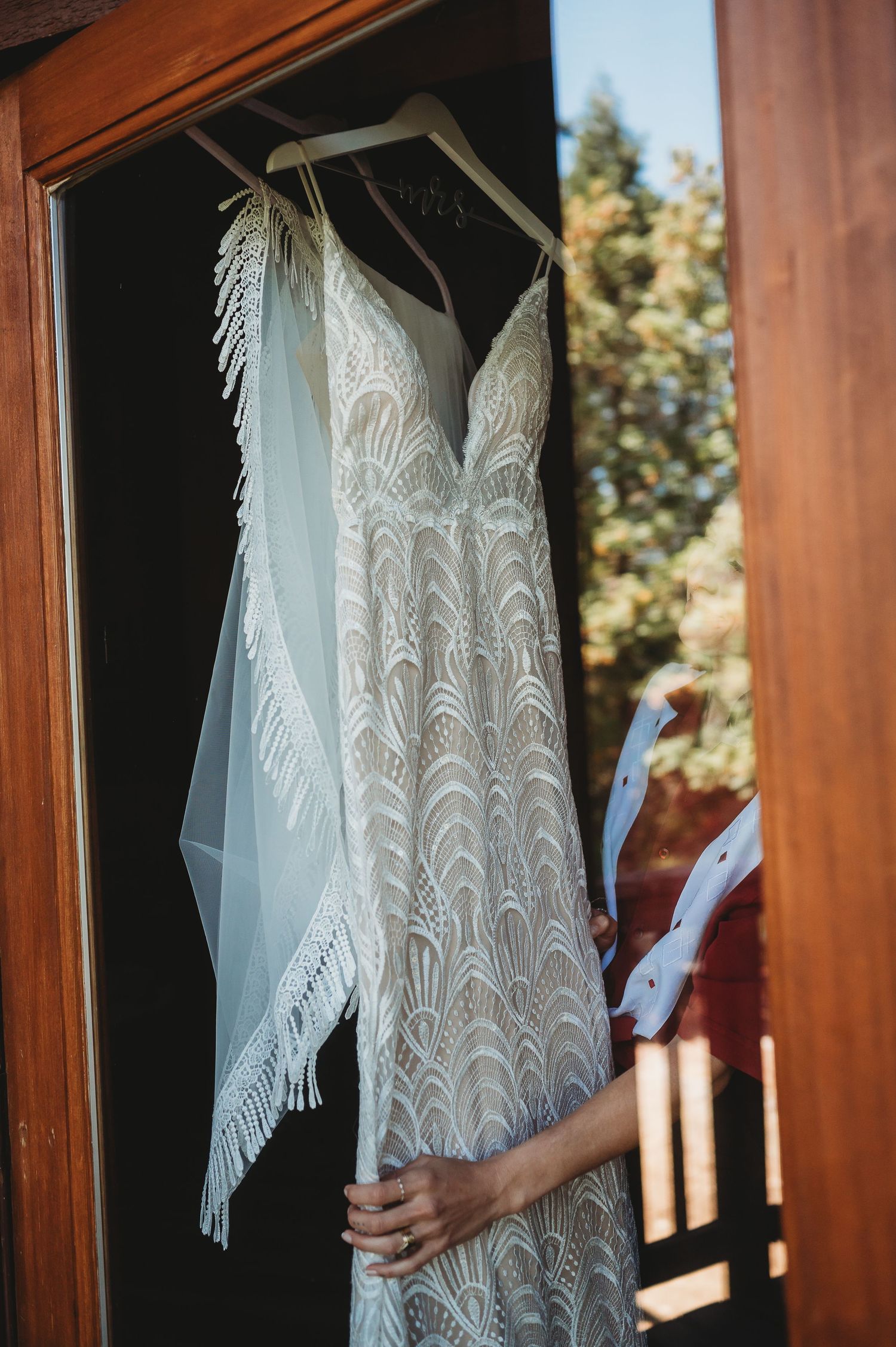 Elegant white lace wedding dress hanging in rustic wood cabin with sunlight streaming through the window.