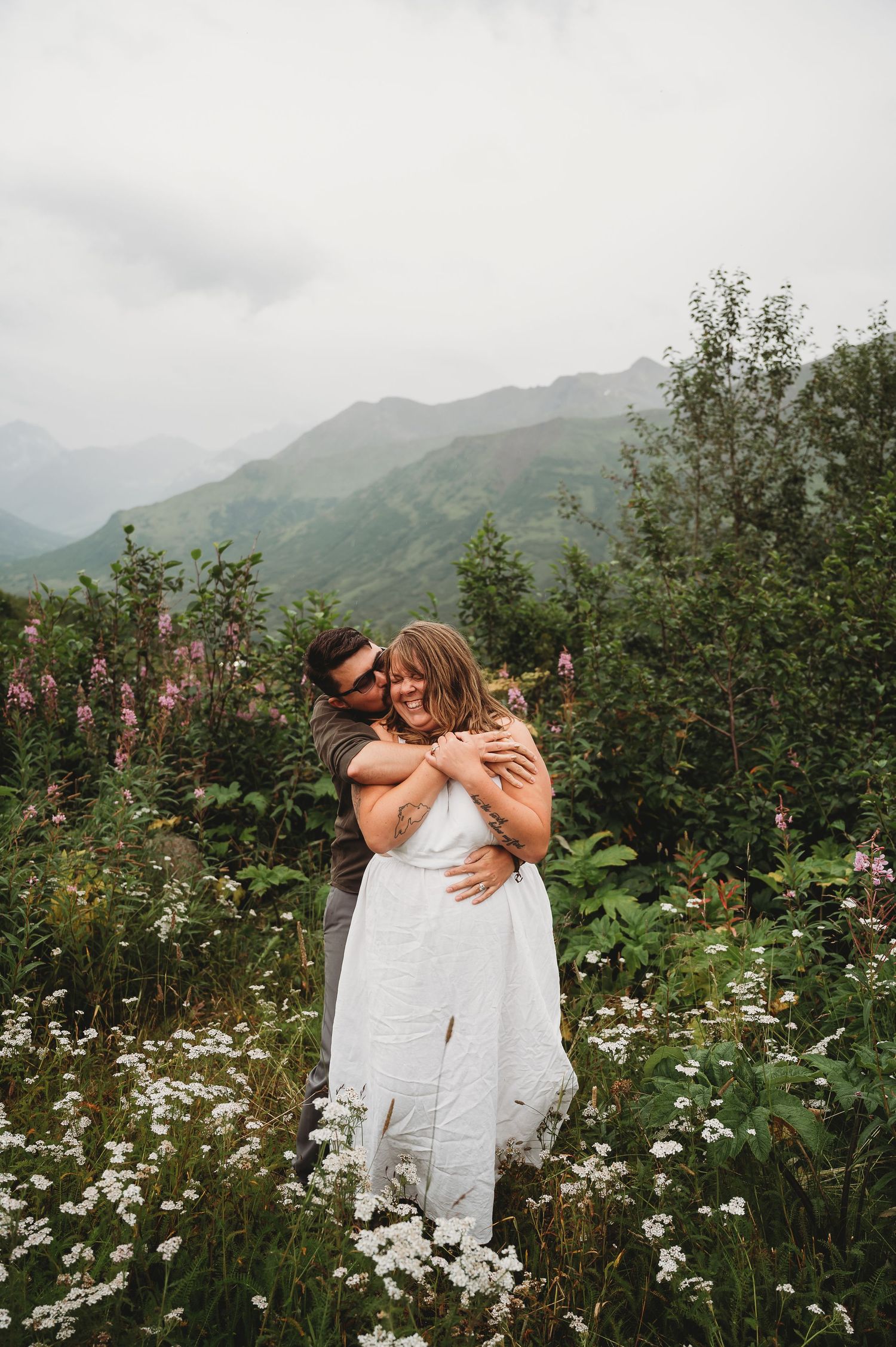 Two people share an intimate moment surrounded by white wildflowers and mountain scenery.