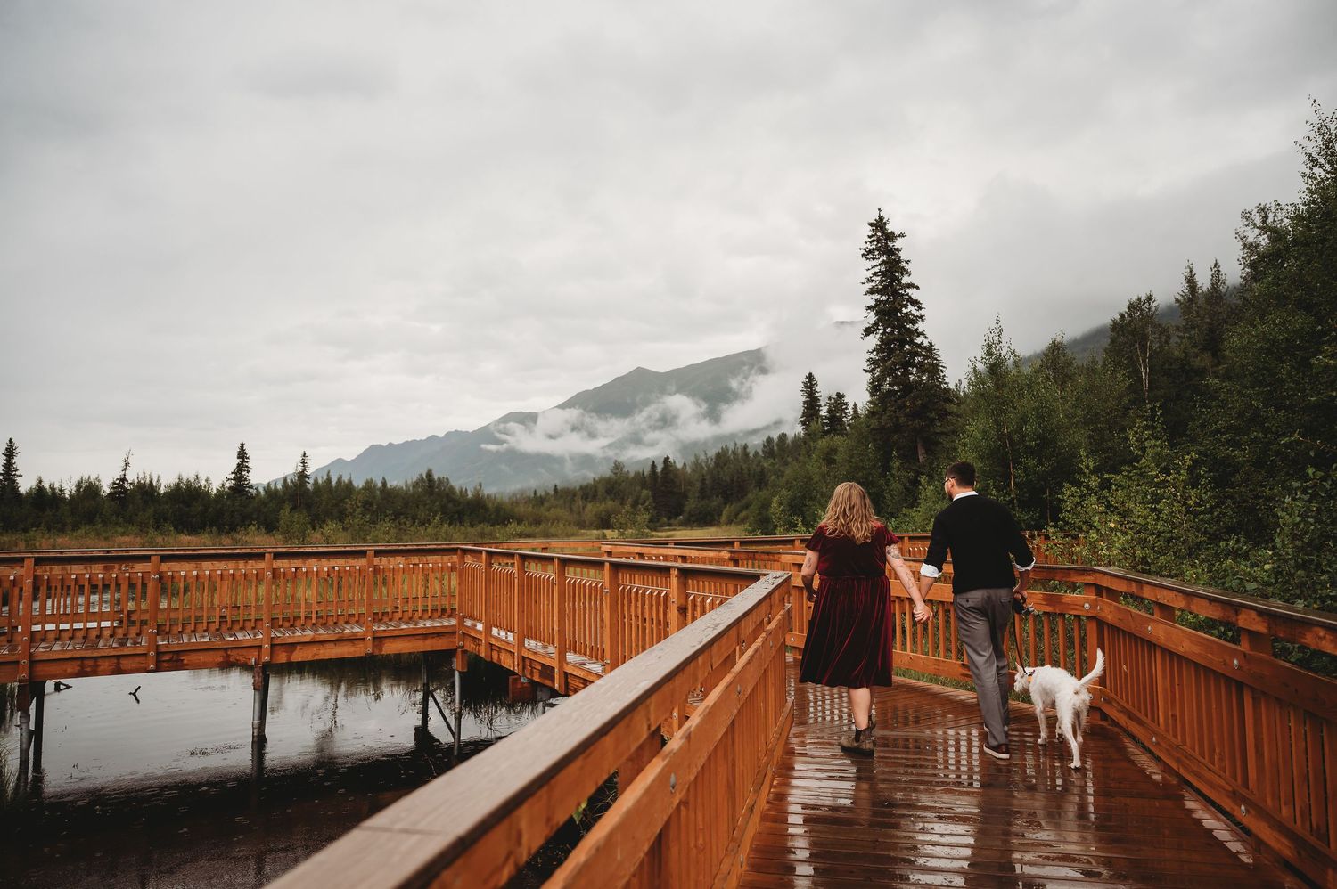 Long wooden boardwalk extends over wetlands with snow-capped mountains and evergreen forest in background.
