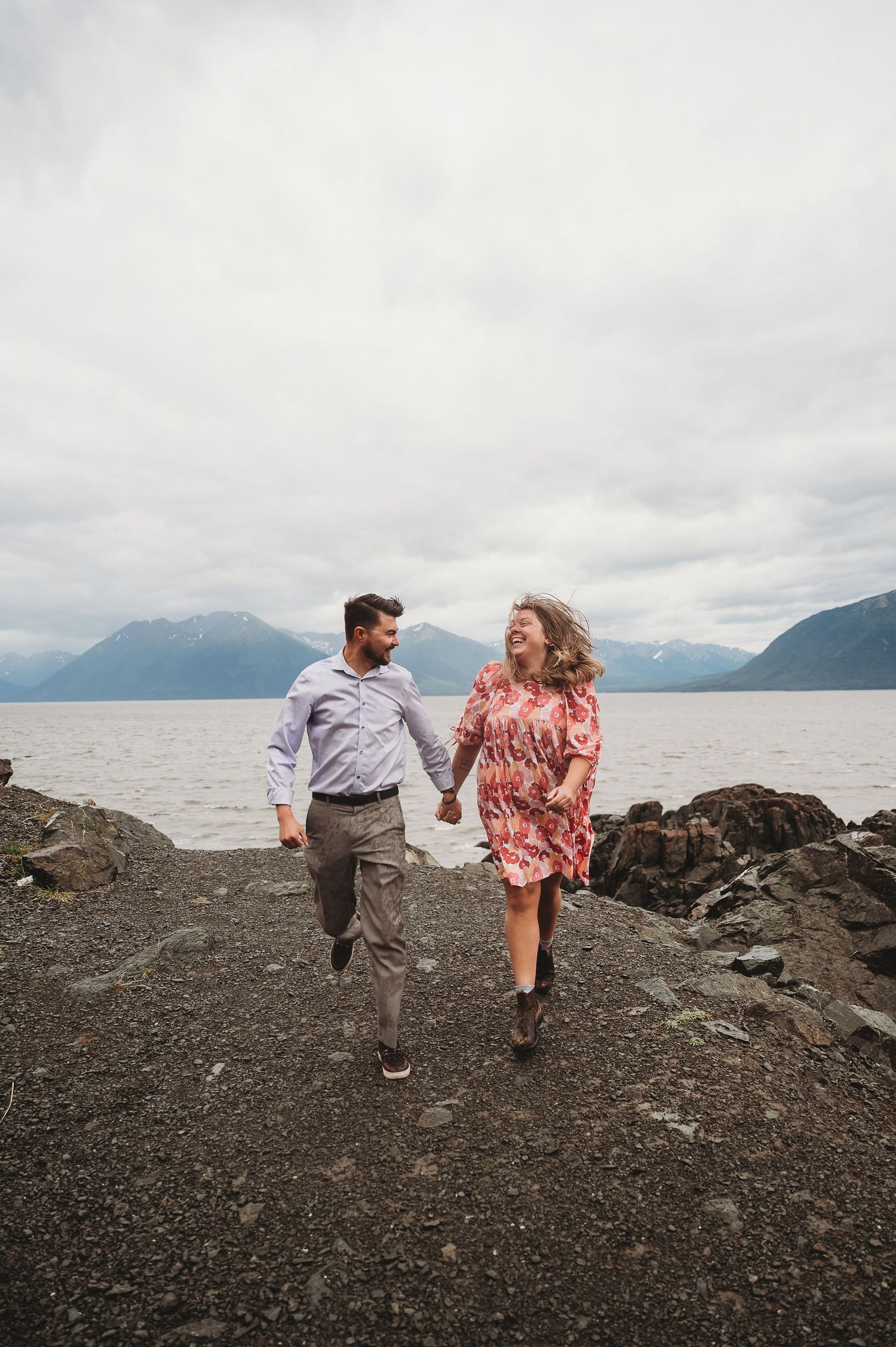 A couple runs hand in hand on a rocky beach with mountains and cloudy skies in the background.