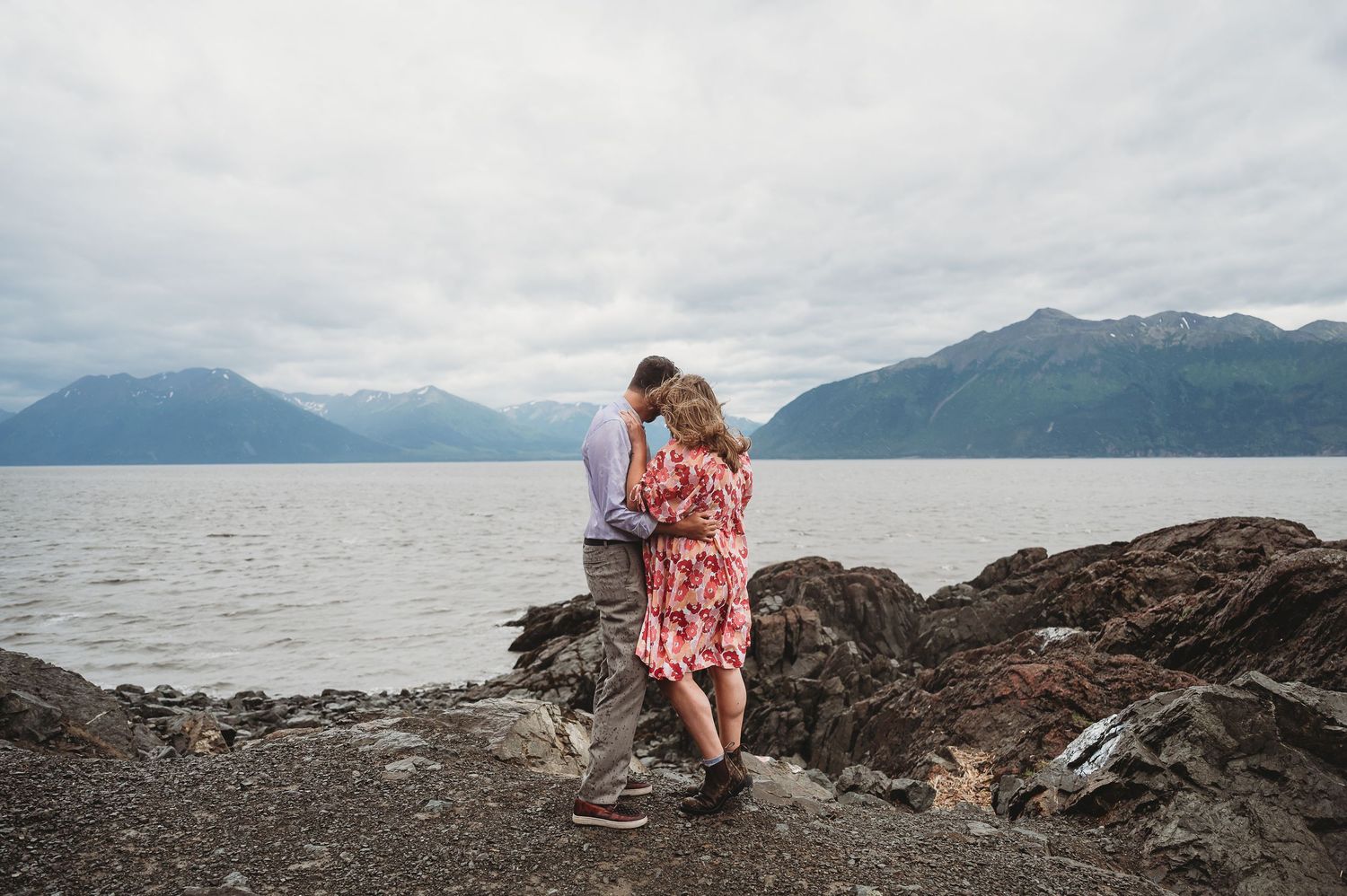 A couple embraces on rocky coastal terrain with mountains and a lake in the background on an overcast day.