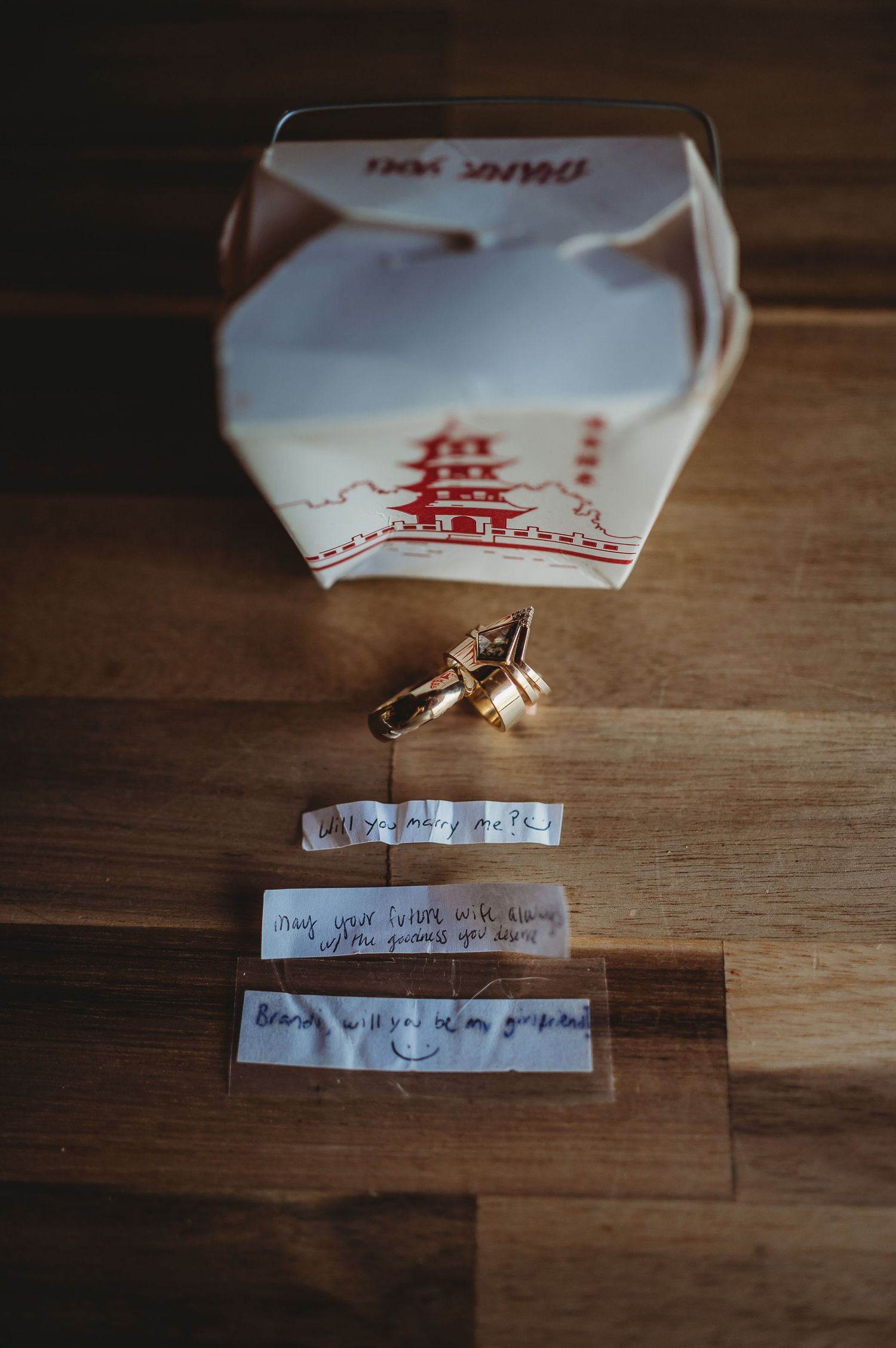 Chinese takeout box with red pagoda design sits beside fortune cookie strips and a ring on wooden surface.