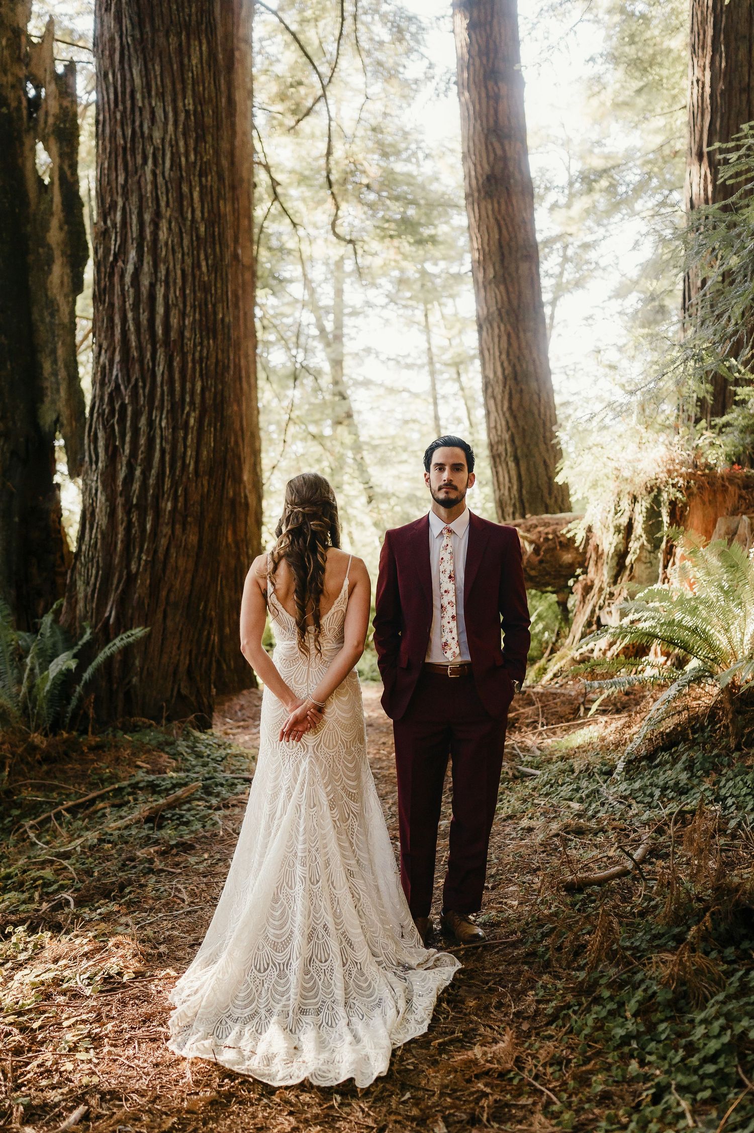 A wedding couple poses dramatically among towering redwood trees on a forest path.
