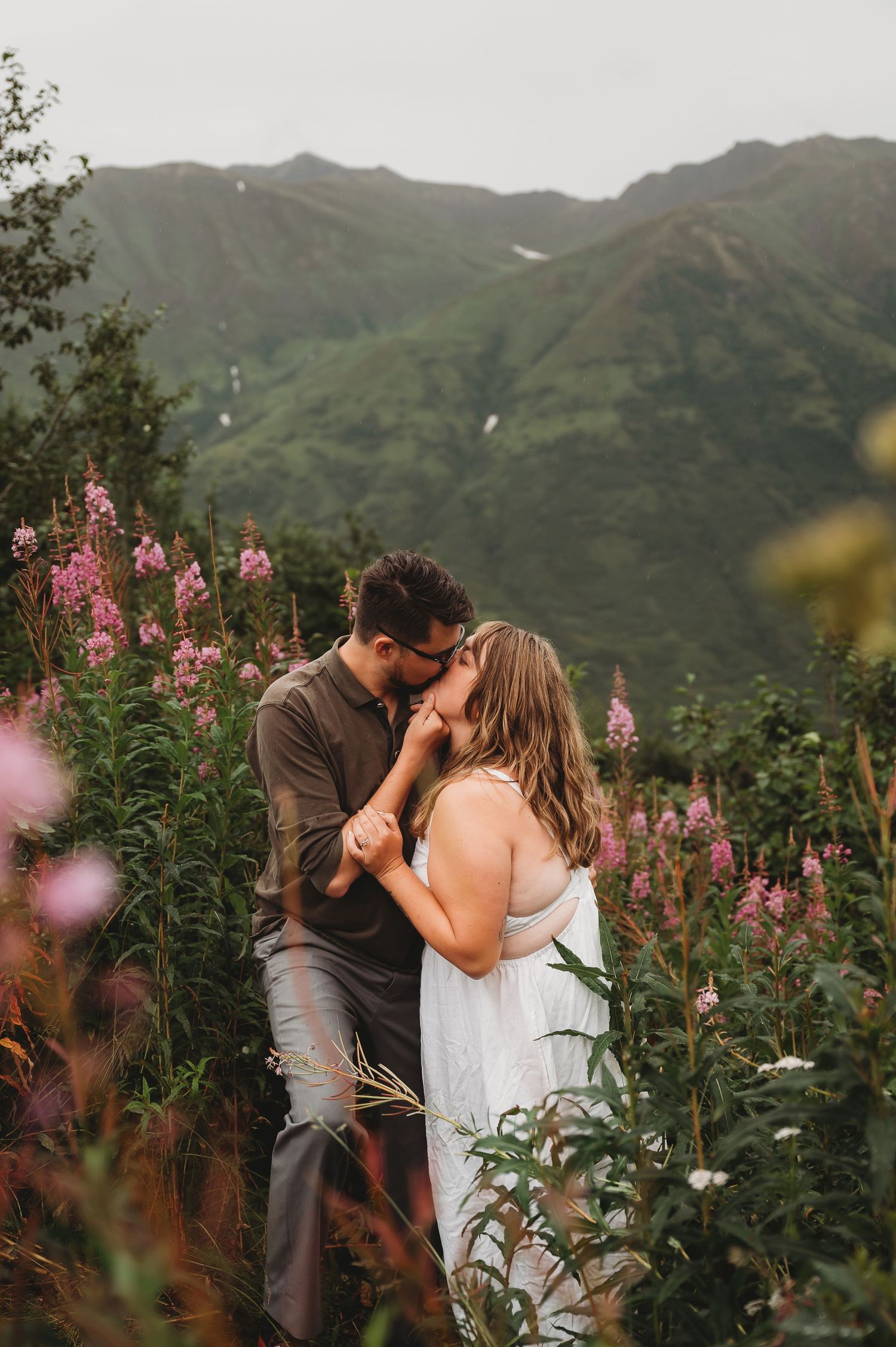 Two people embrace in a mountainside meadow filled with pink fireweed flowers at sunset.