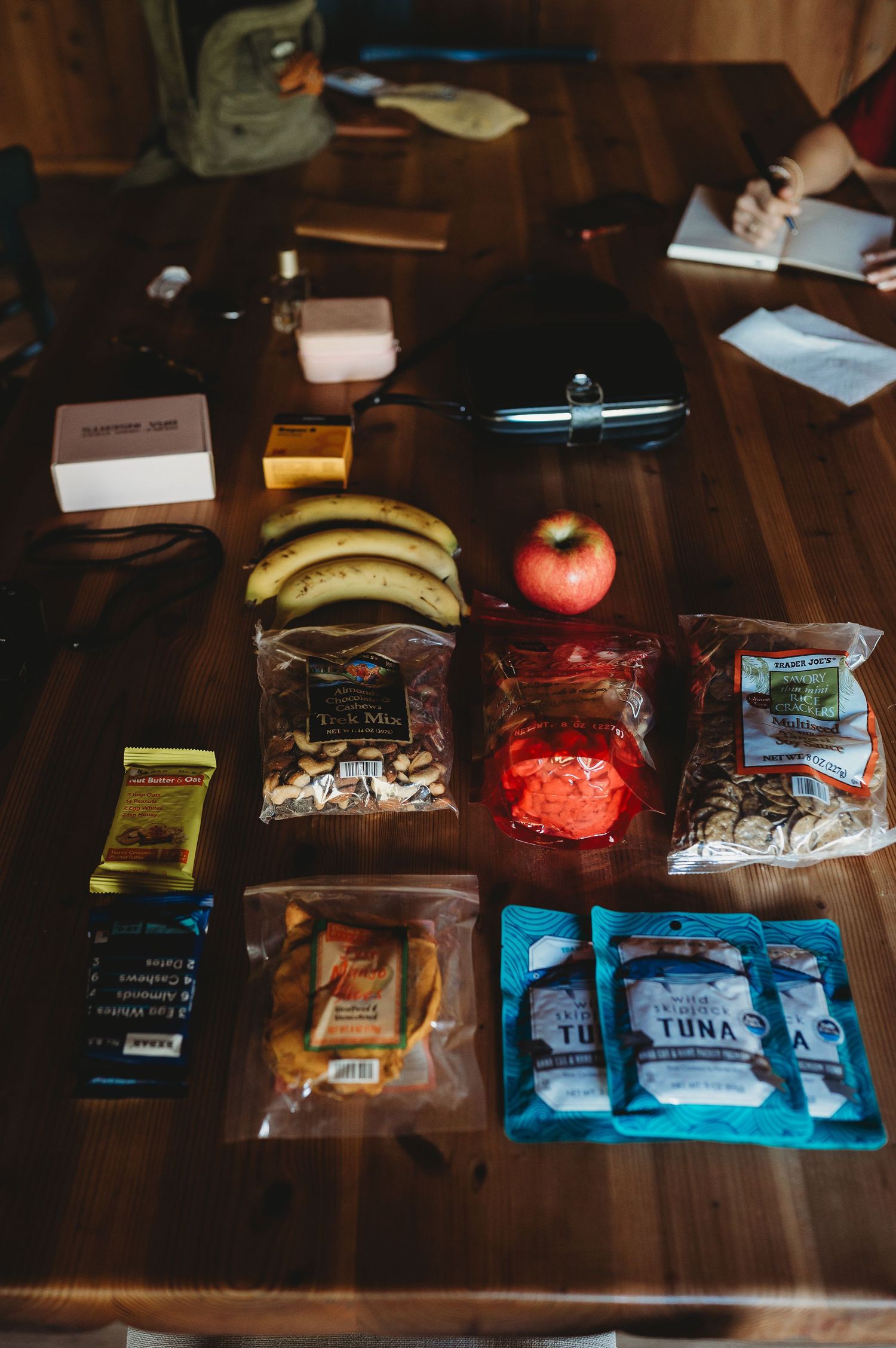 Travel snacks and food items laid out on wooden table including fruit, tuna, and packaged goods.