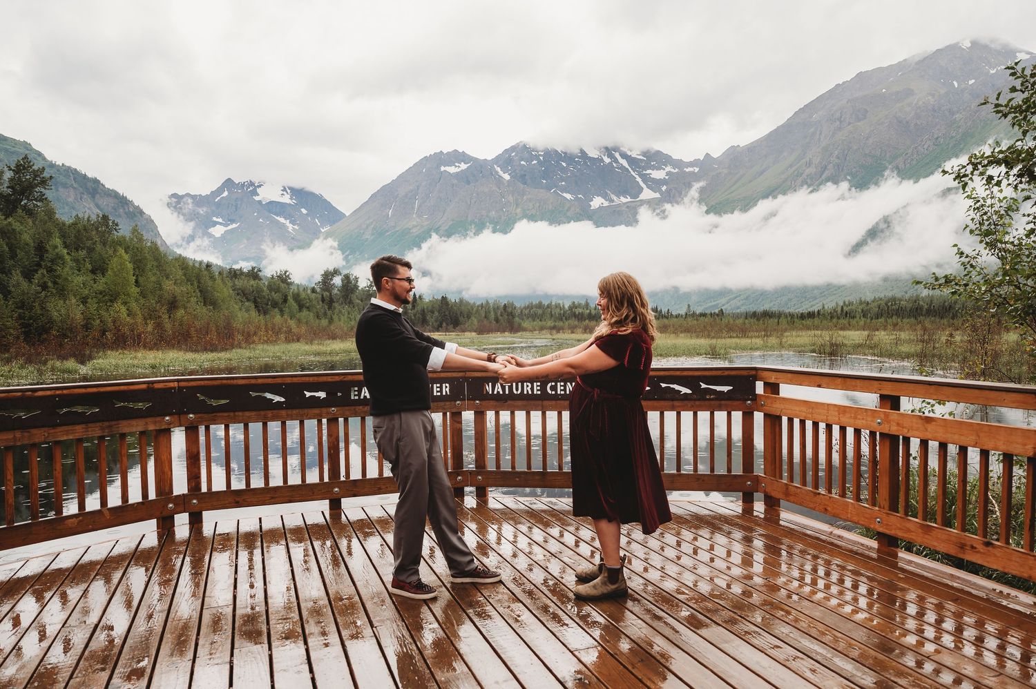 Couple on a wooden observation deck overlooking snow-capped mountains and misty valleys.