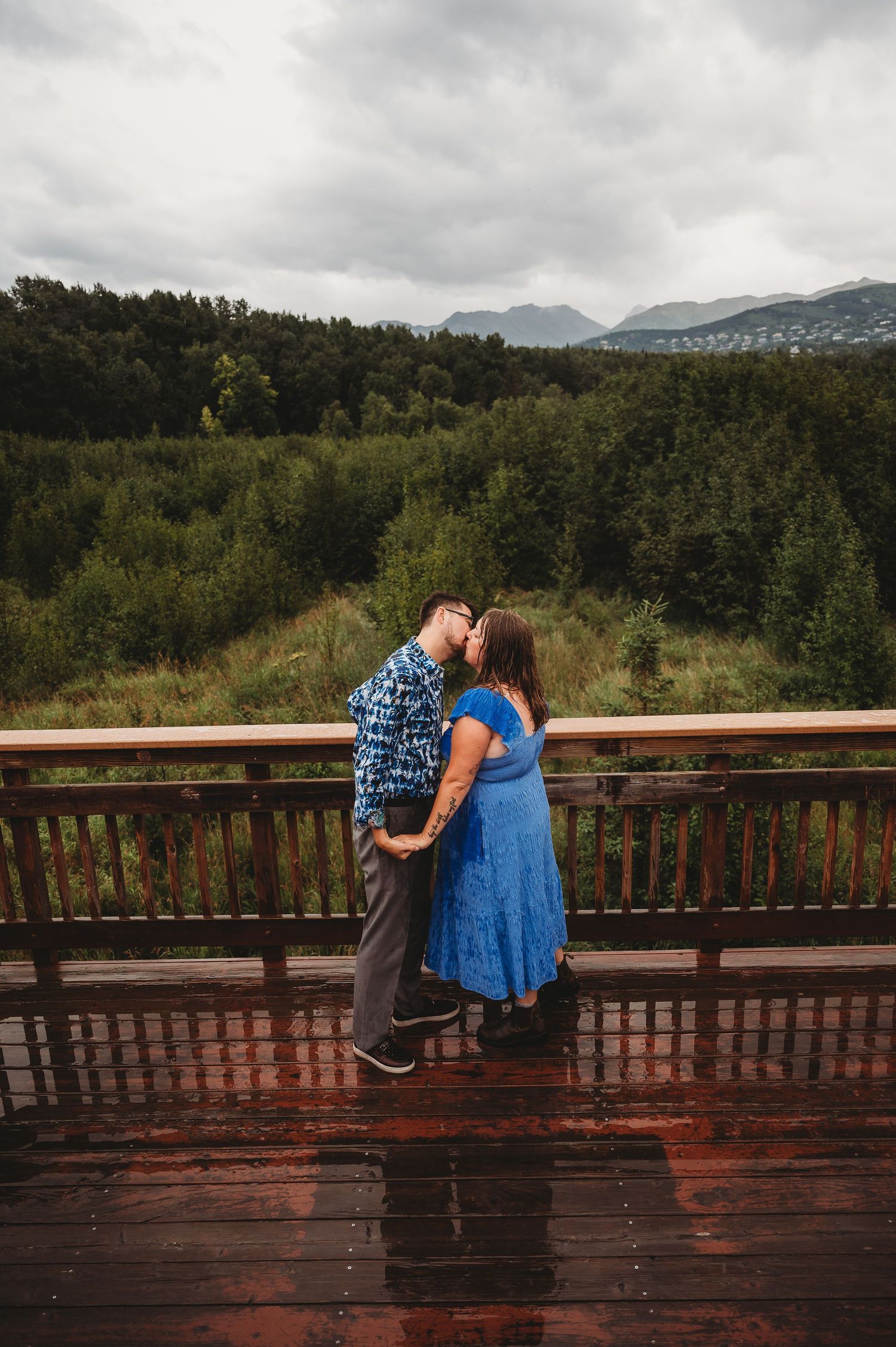 A romantic moment shared on a wet wooden deck overlooking a mountainous forest landscape on a cloudy day.