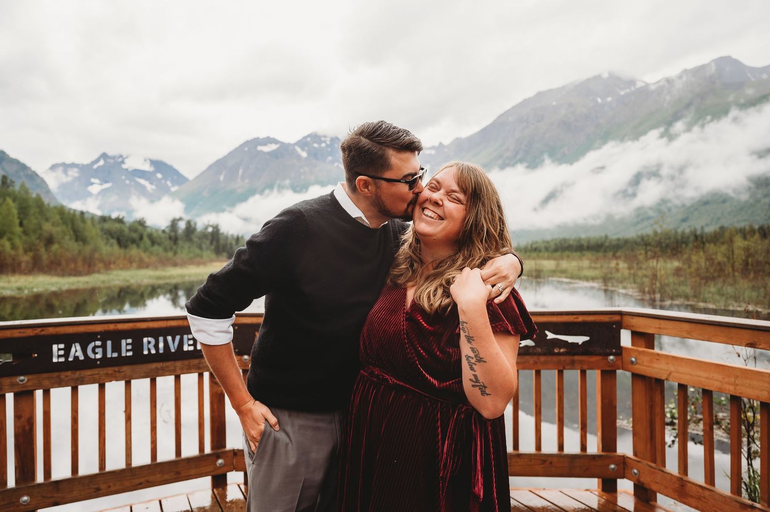 Joyful moment shared between couple laughing on viewing platform with misty Alaskan mountain landscape behind them.