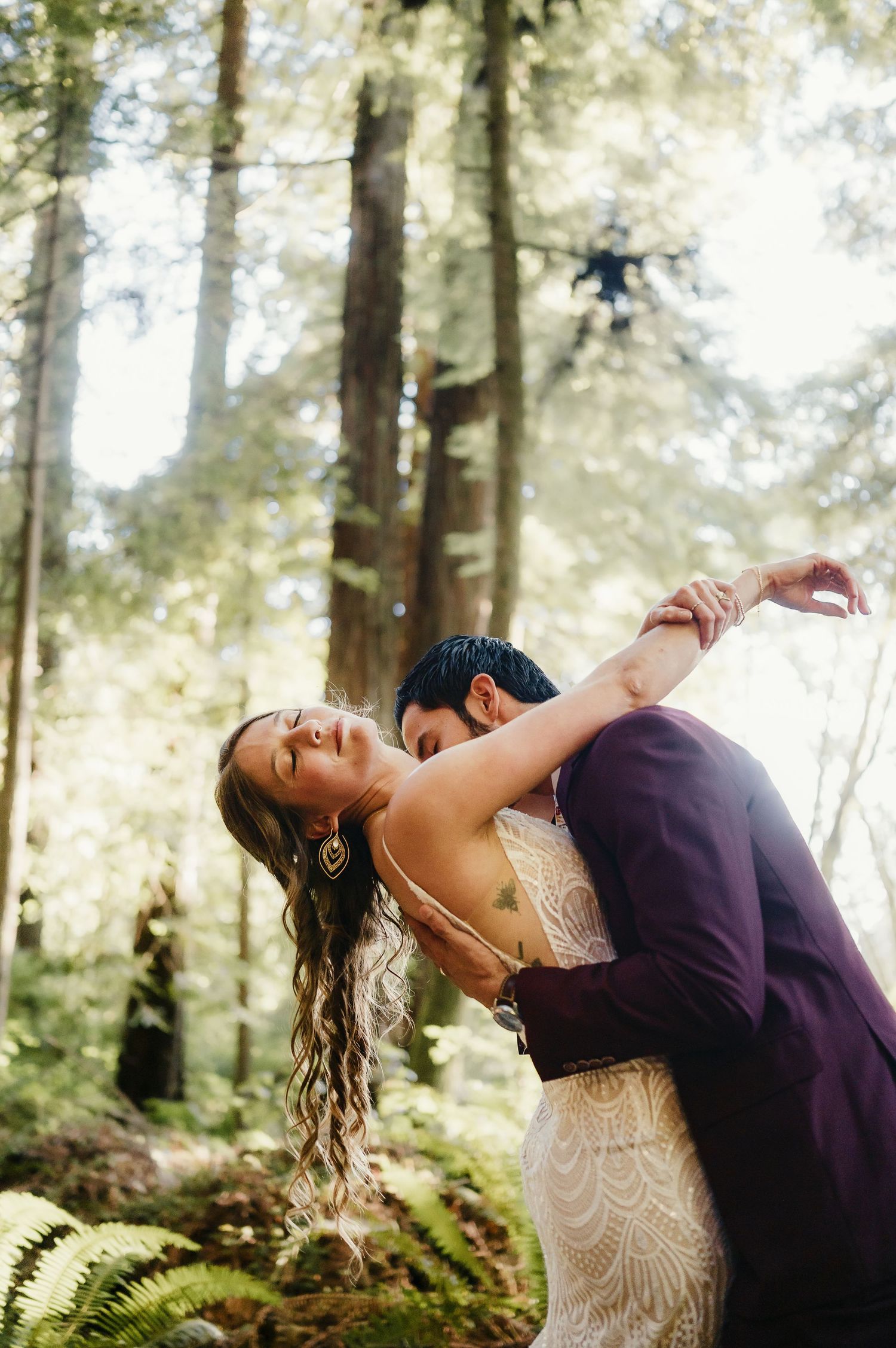 Romantic dip pose captured in a sunlit forest with ferns and towering trees.