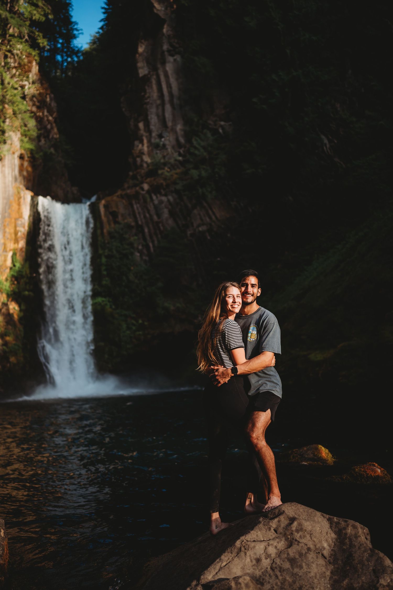 A couple enjoys a romantic moment near a scenic waterfall in a lush forest setting during golden hour.