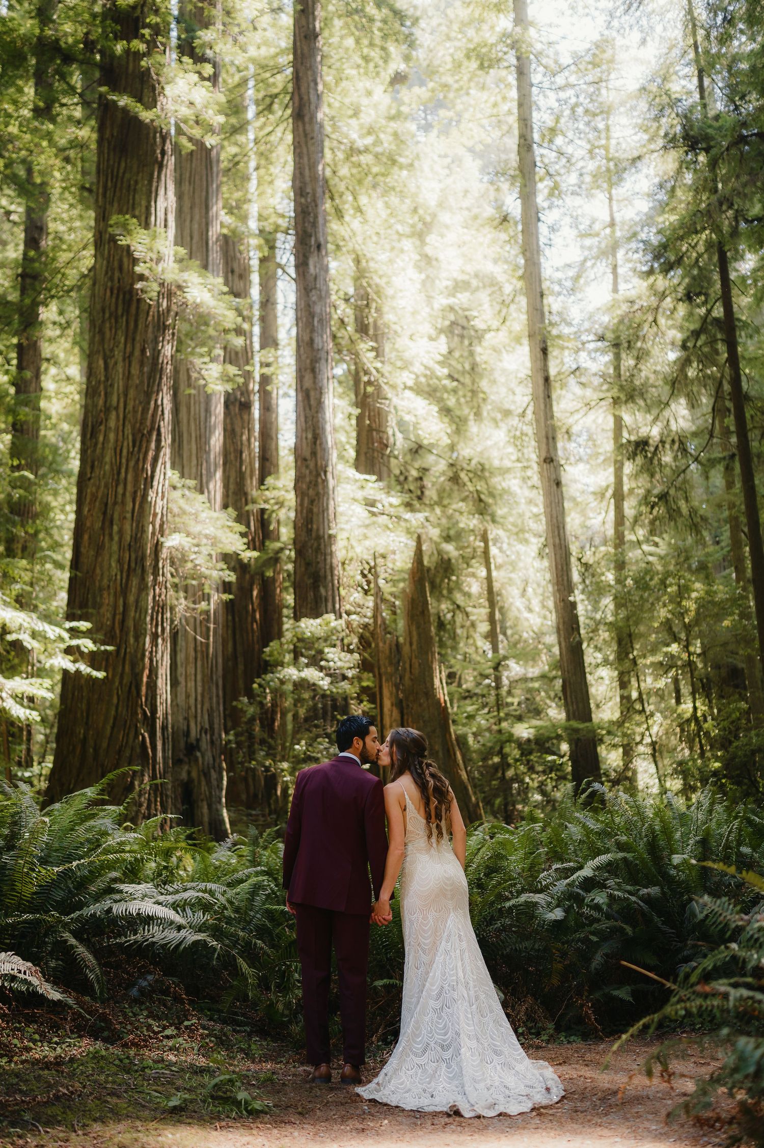 A couple poses in a dreamy redwood forest setting surrounded by towering trees and lush ferns in dappled sunlight.