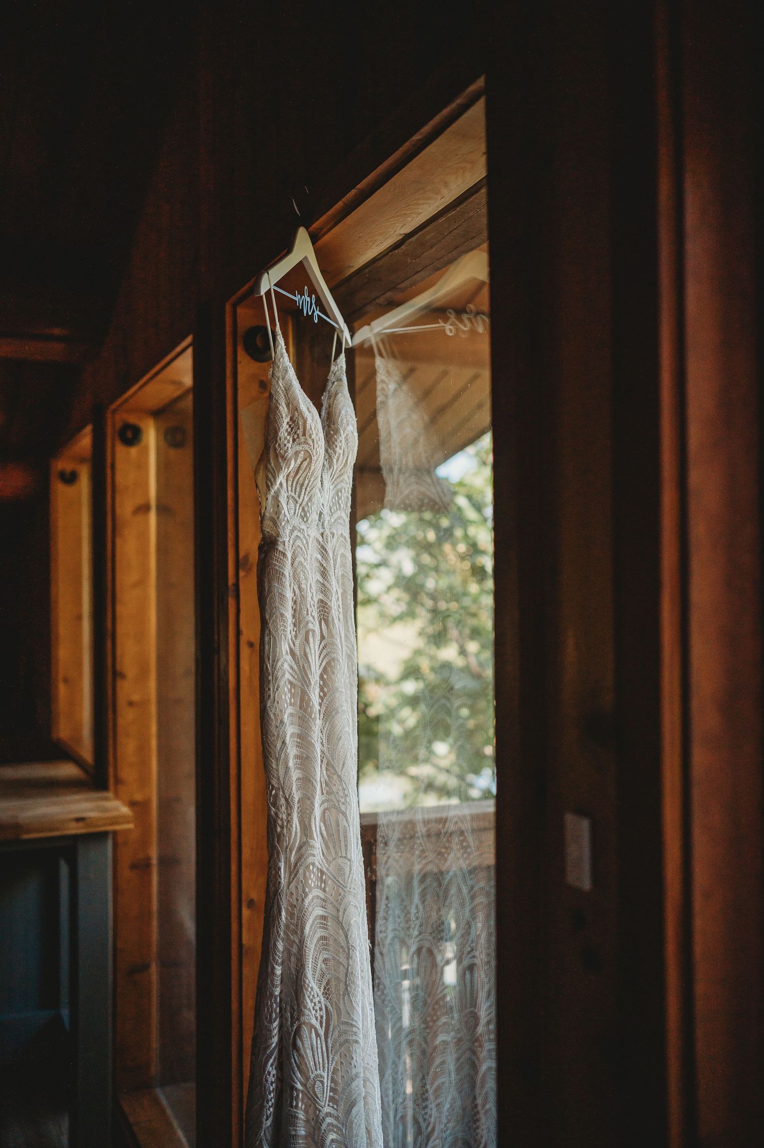 White lace wedding dress hanging in sunlit window of wooden cabin.